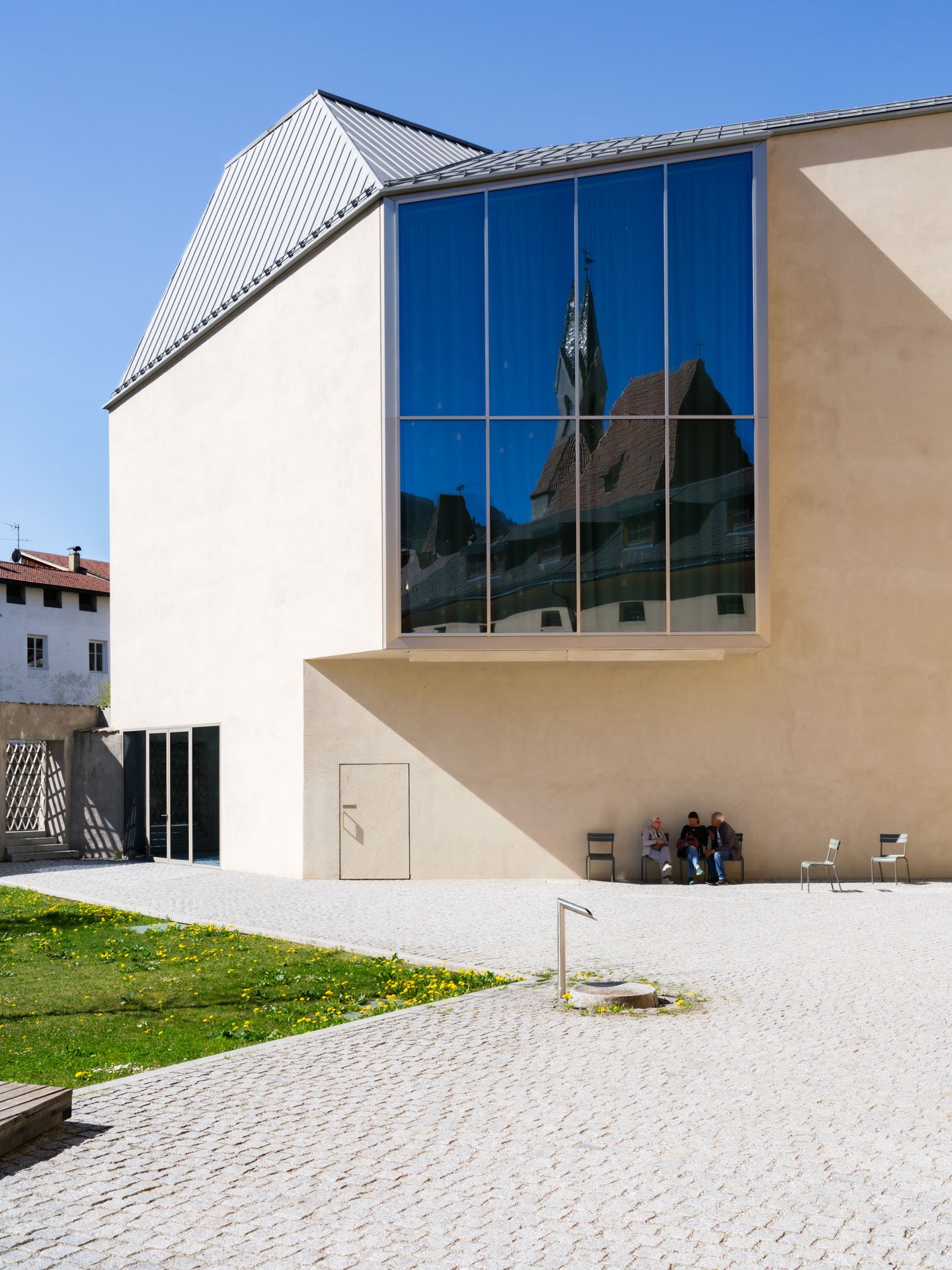 Una moderna costruzione con una grande vetrata che riflette una chiesa storica con un campanile. Sullo sfondo, un cielo blu e alcuni edifici. In primo piano, tre persone sedute su una panchina. L'area circostante è un cortile con pavimentazione in pi