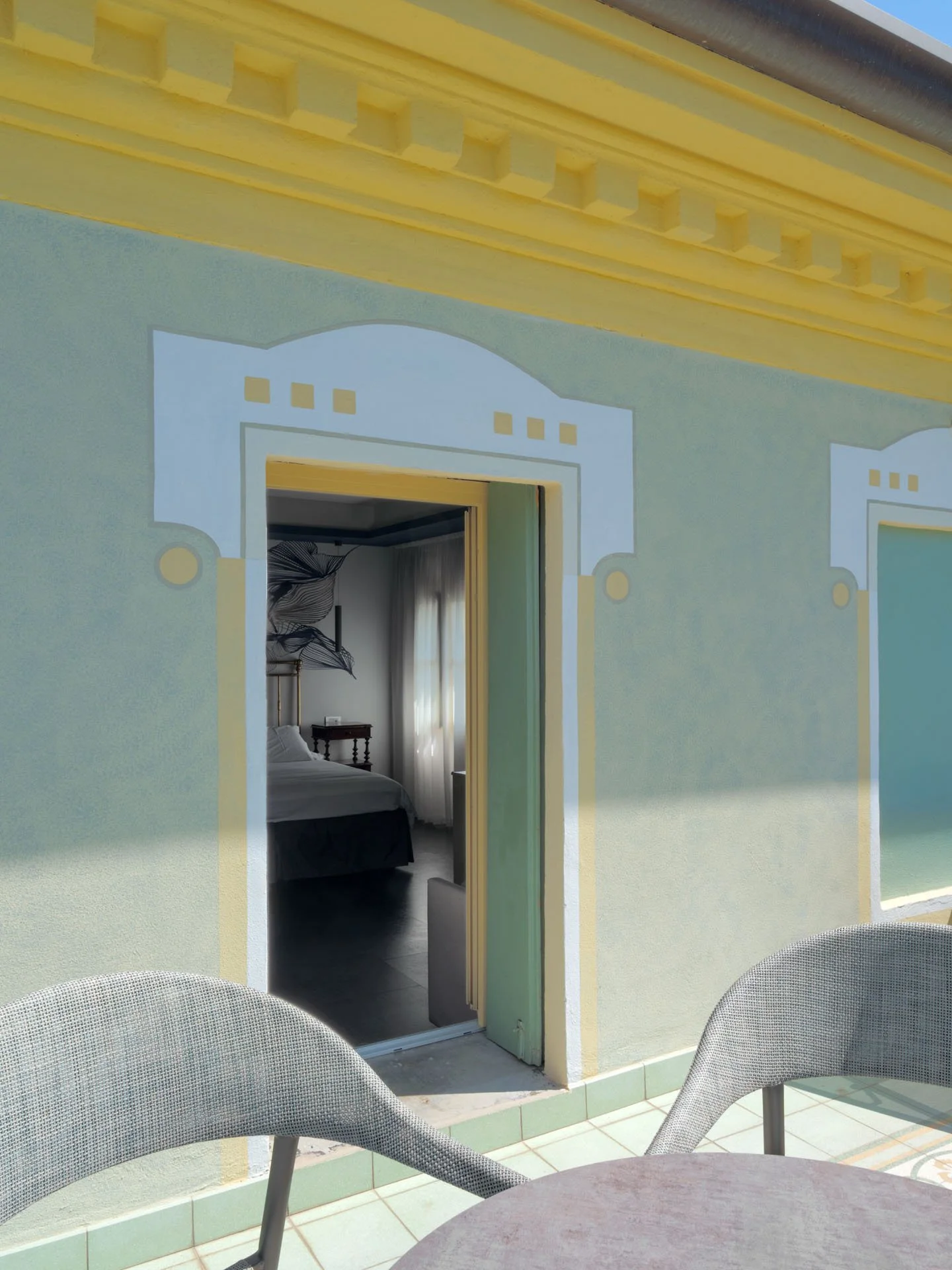 Vista da una terrazza con tavolo rotondo e sedie in plastica, affaccio su una parete colorata con decorazioni ingrandite di porte e finestre, visibile anche una camera da letto interna.