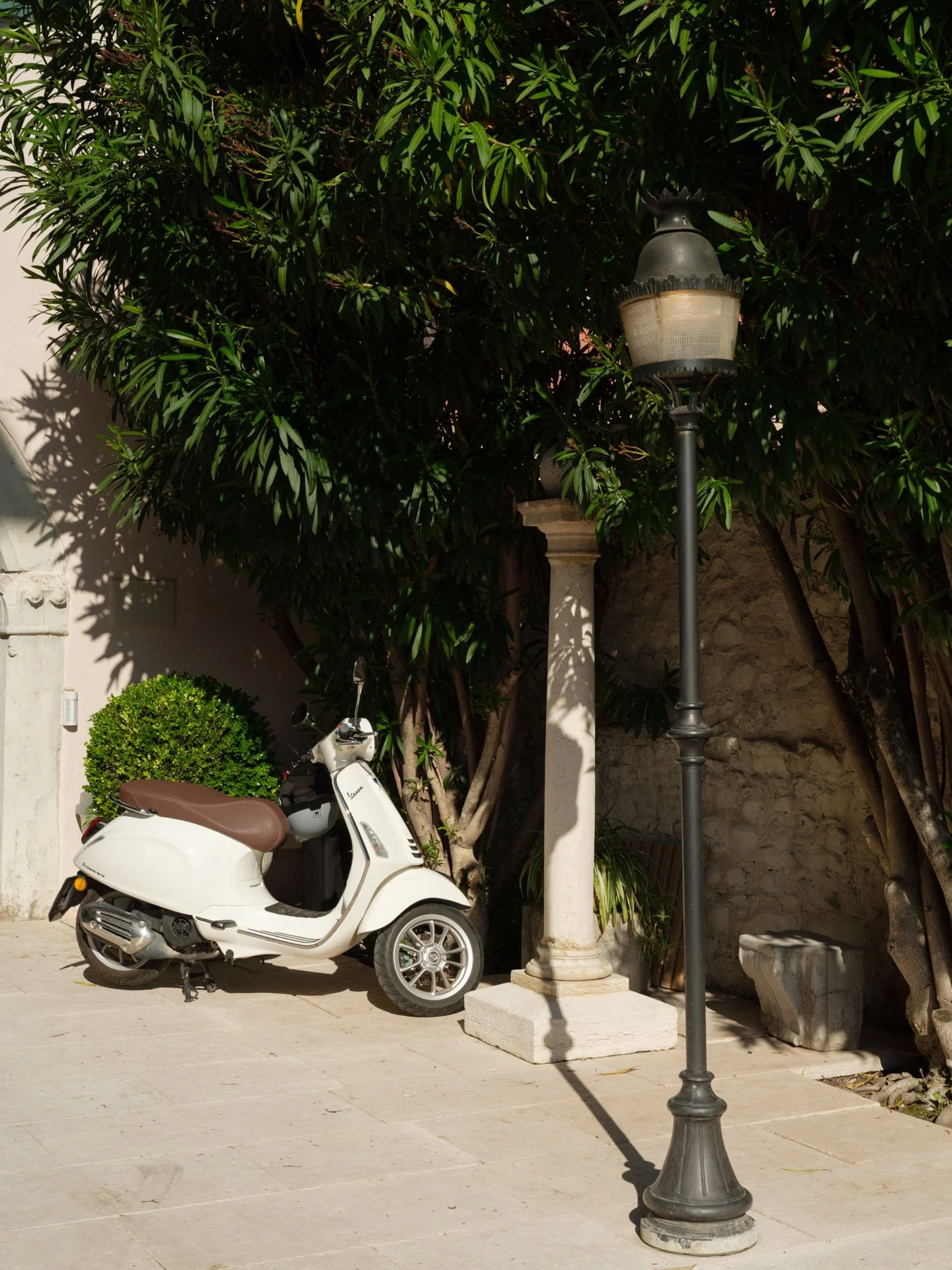 Una scooter bianca parcheggiata vicino a una pianta e una colonna in pietra con un lampione moderno, su una superficie di pietra chiara, con un muro di pietra sullo sfondo.
