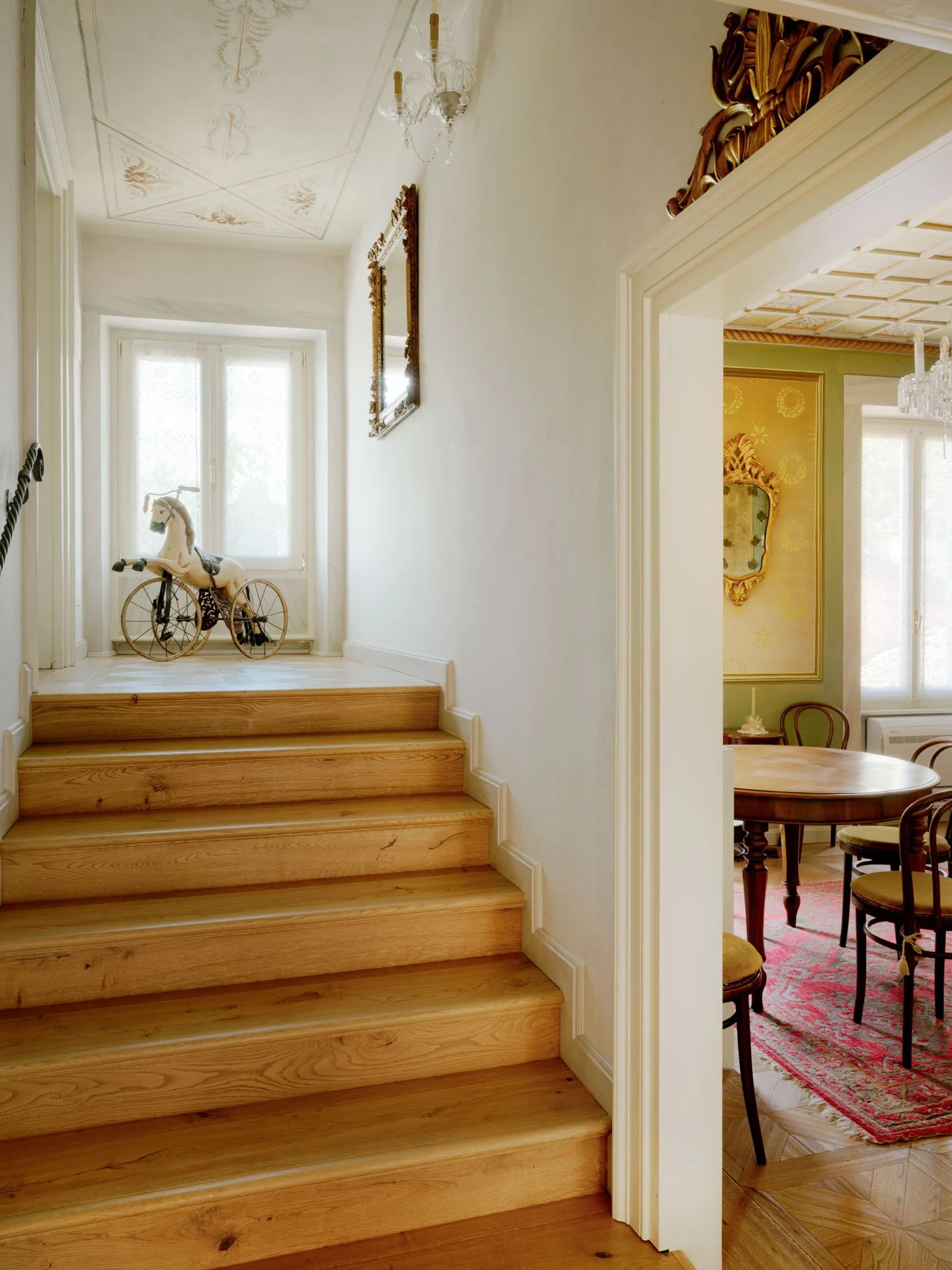 Scala in legno che conduce a un soggiorno con pareti bianche, decorazioni classiche, vicino a un vischio e uno specchio dorato, con una zona pranzo con tappeto rosso e sedie in legno.
