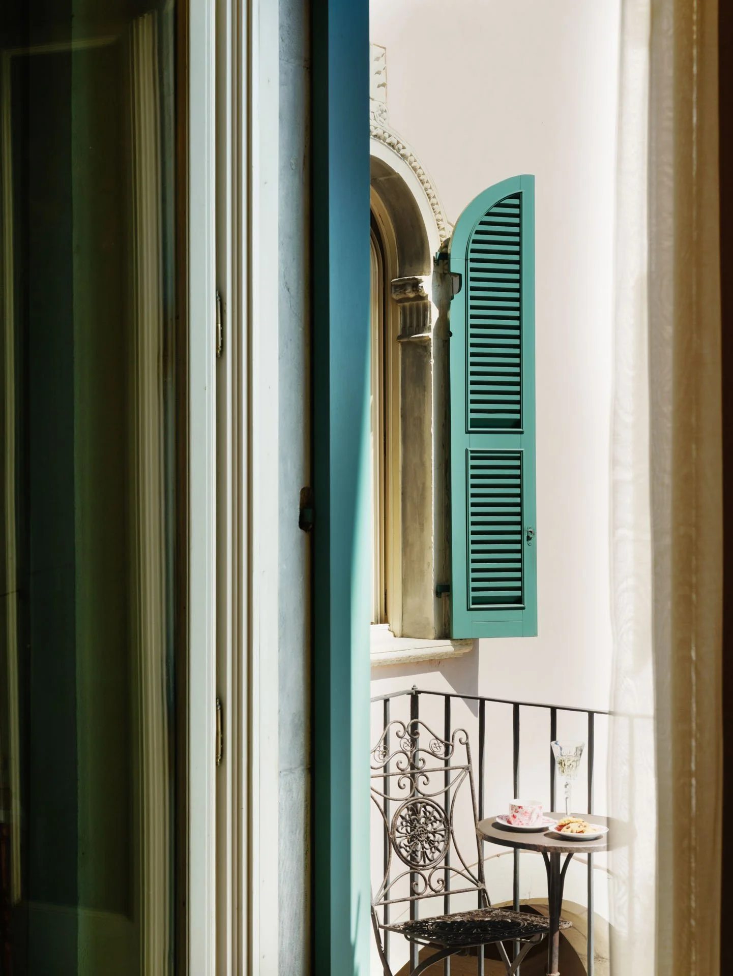 Vista di un balcone attraverso una finestra, con una sedia in ferro battuto, un tavolino con tazze e piatti di cibo, e una finestra con tenda leggero.