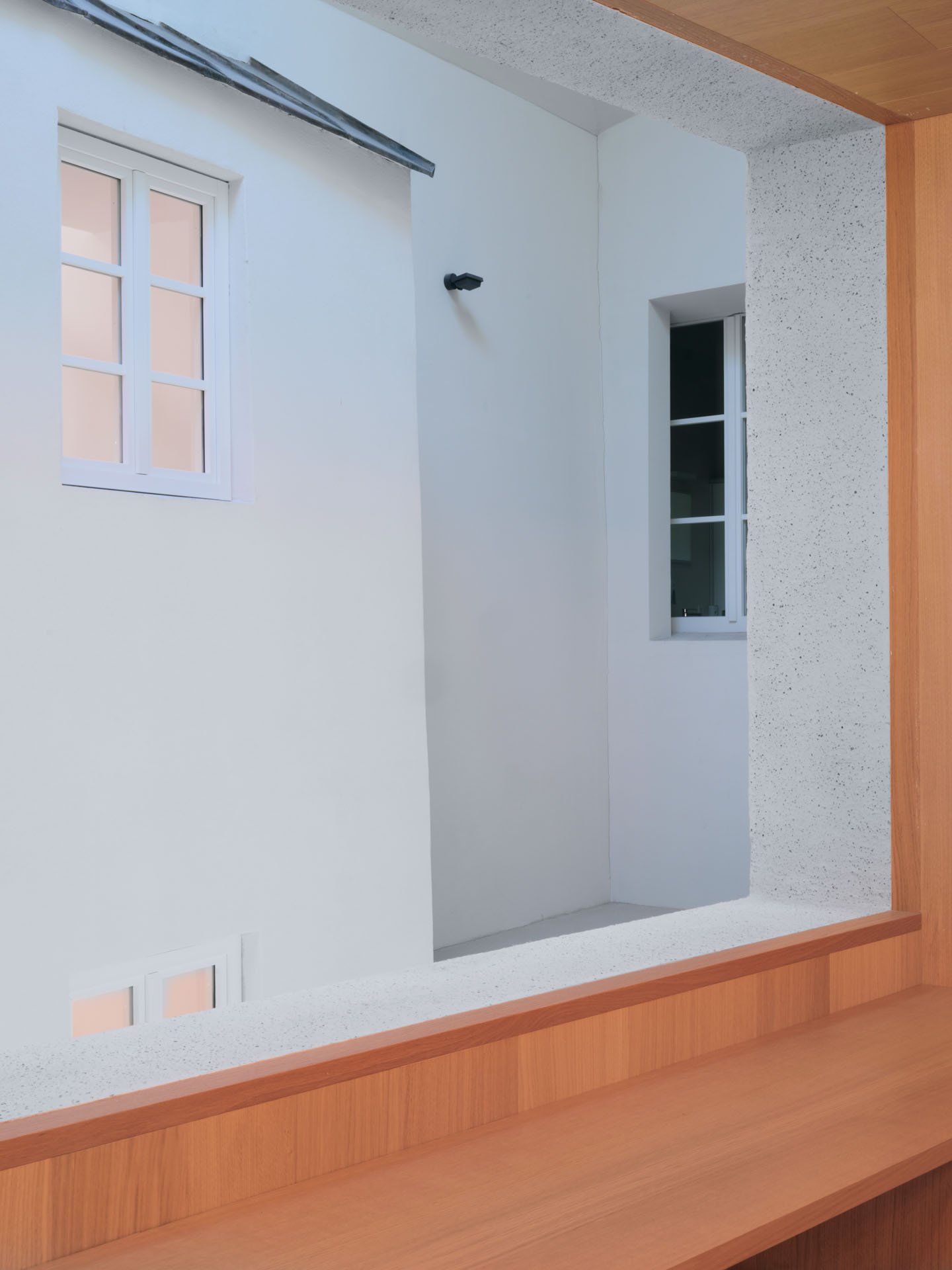 Interno di un edificio con pareti bianche, due finestre con telai bianchi, una parete rivestita in legno e un soffitto in cemento con una tubazione metallica.