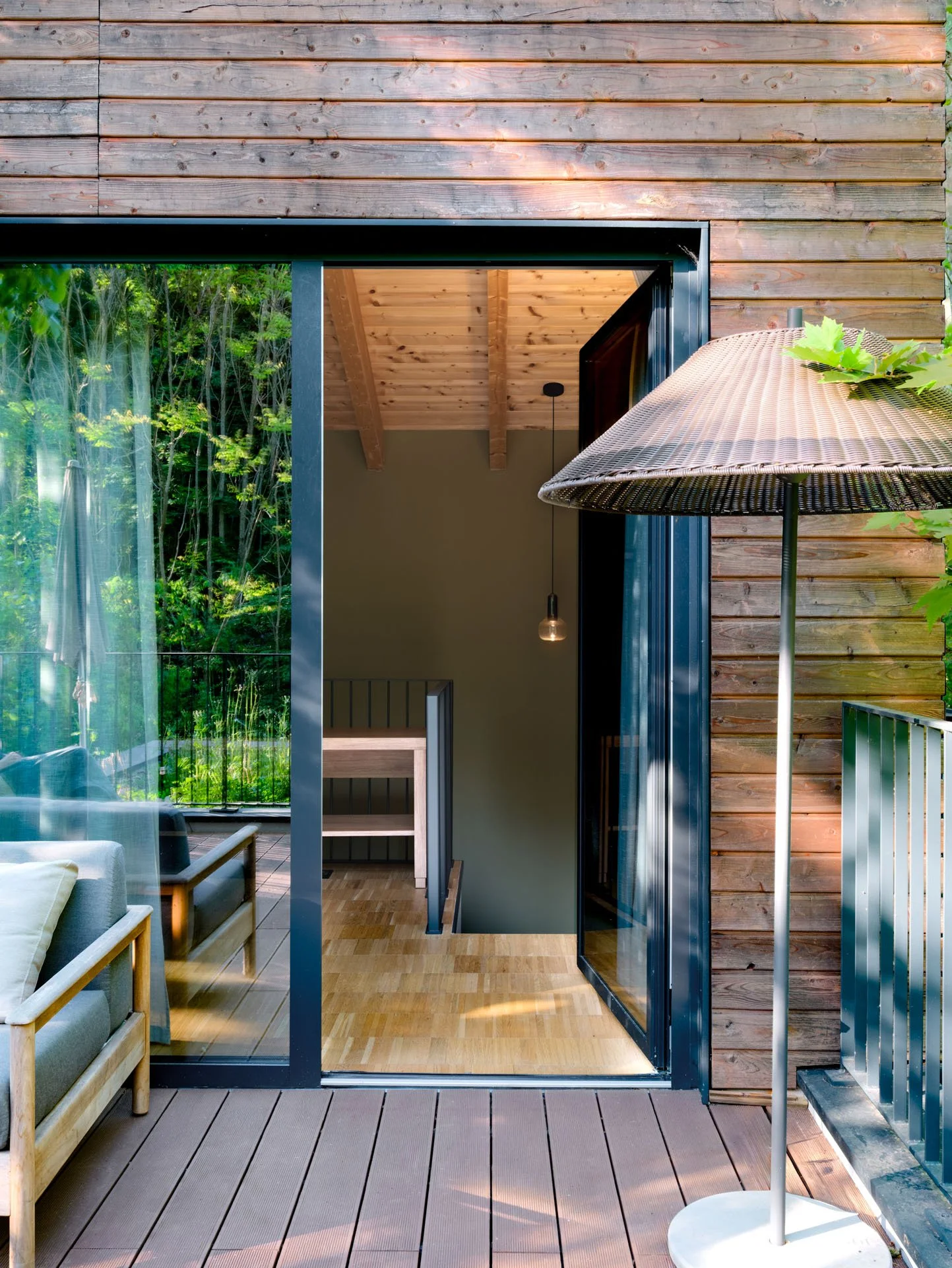 Foto di una terrazza in legno con un grande ombrellone, una porta aperta che conduce a una stanza con soffitto in legno e una finestra grande che mostra una vista di alberi verdi.