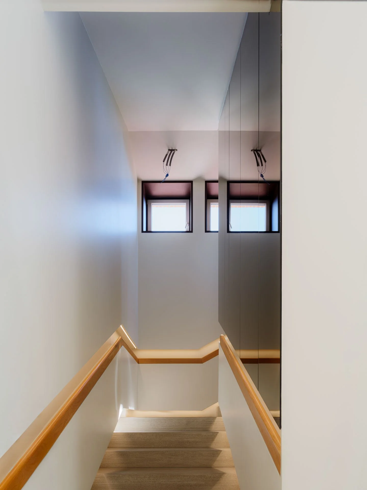 Scalinata in un interno con pareti bianche, corrimano in legno, debole illuminazione e due finestrini sopra che lasciano passare luce naturale.