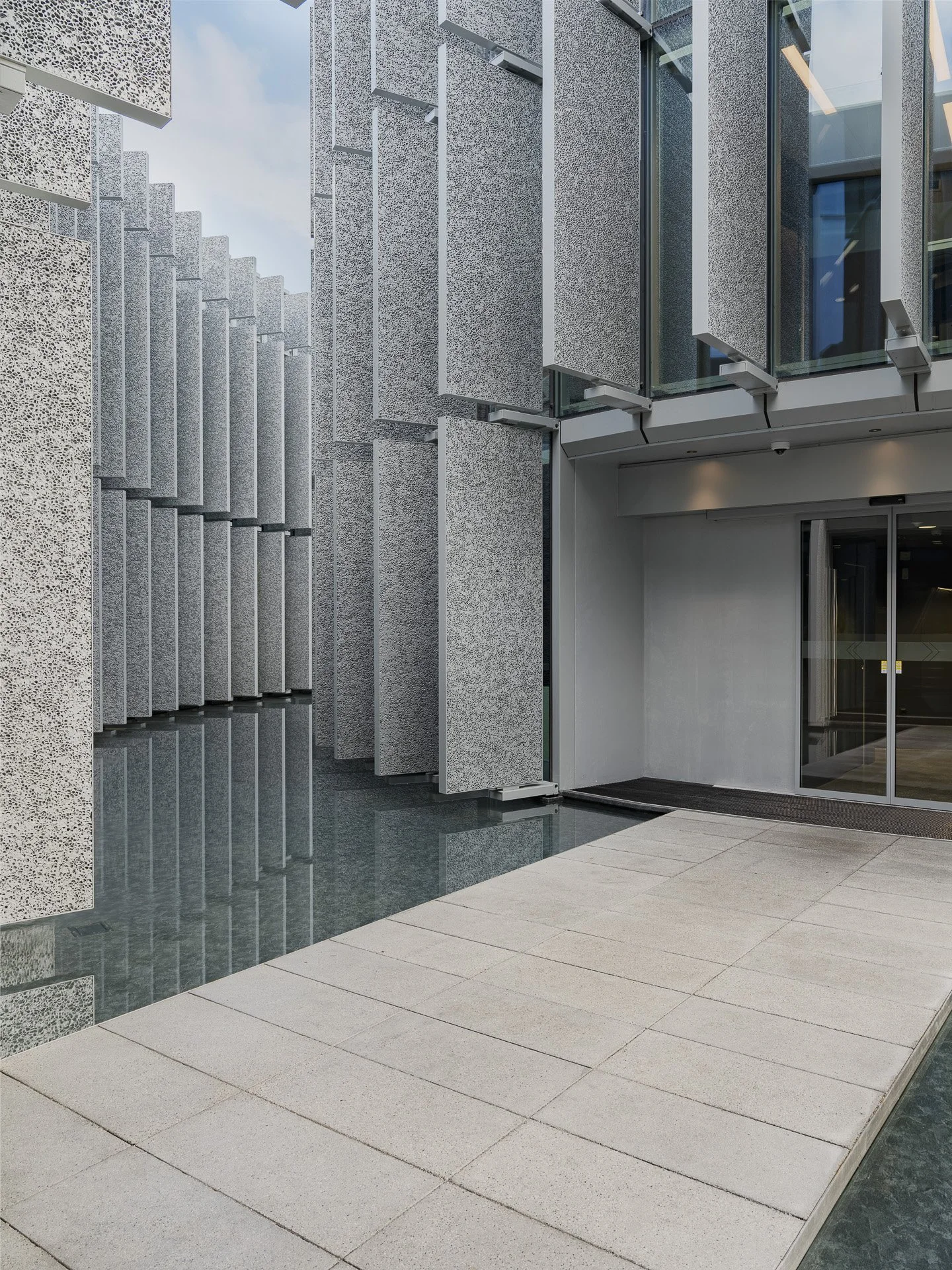 Esterno di un edificio moderno con pareti in pietra e dettagli in vetro, ingresso con porta scorrevole e pavimentazione in piastrelle