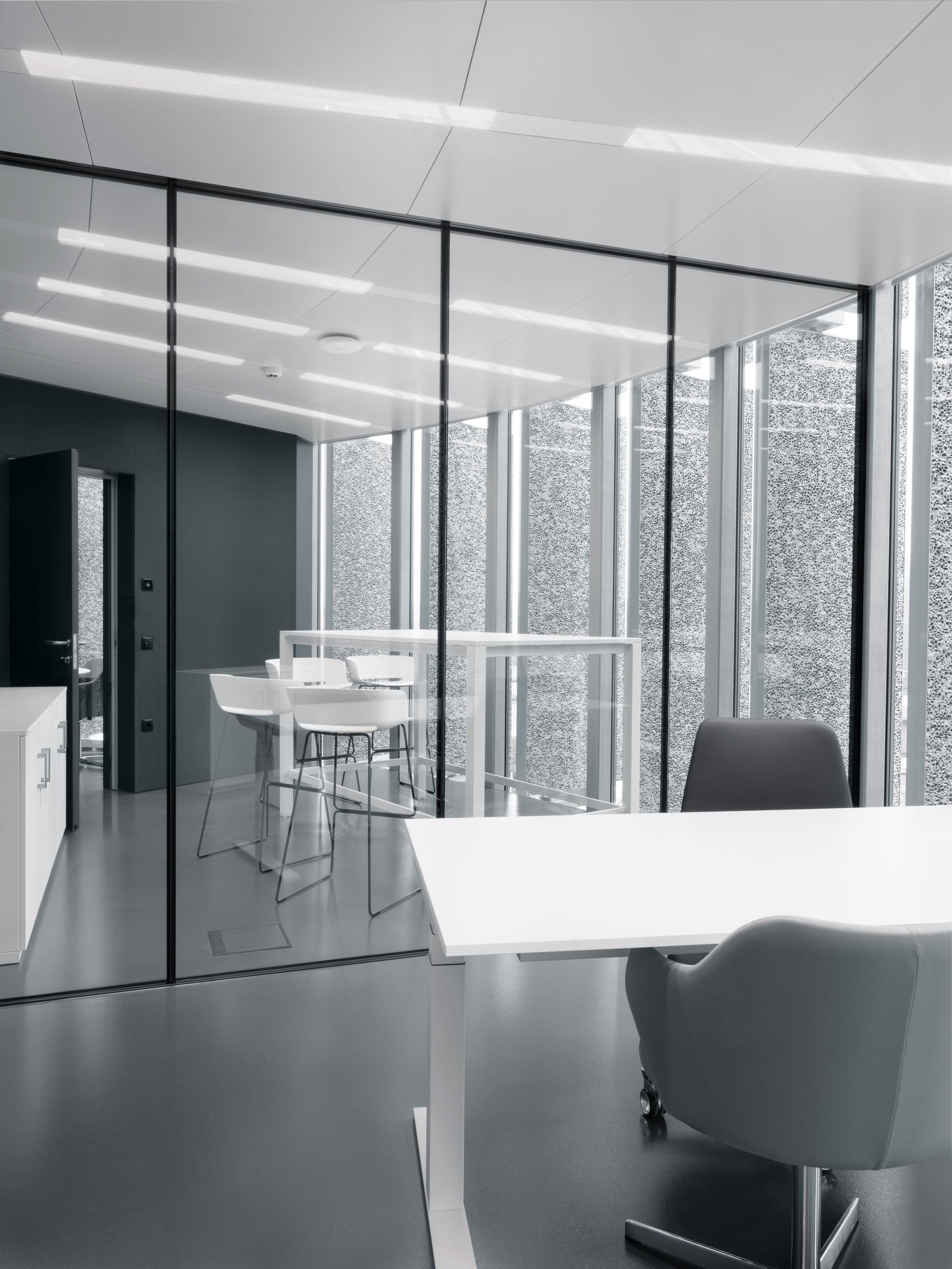 Vista di un ufficio moderno con pareti in vetro, scrivanie bianche, sedie bianche e una parete di retro con un motivo decorativo. Ambiente luminoso e minimalista.