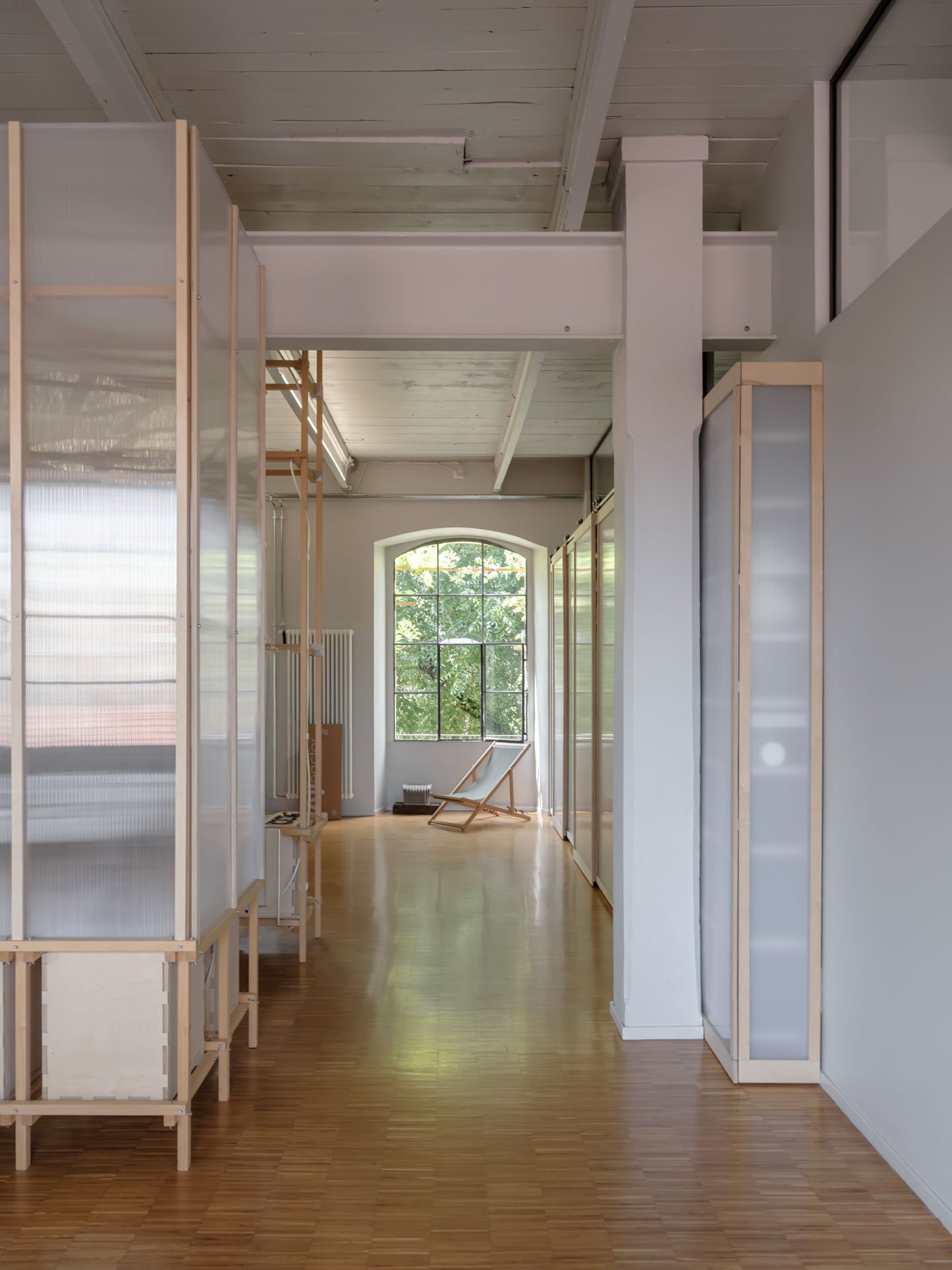Interno di uno spazio moderno con scaffali in legno e pareti bianche, grandi finestre che lasciano entrare luce naturale, e una sedia a sdraio di legno vicino alla finestra.