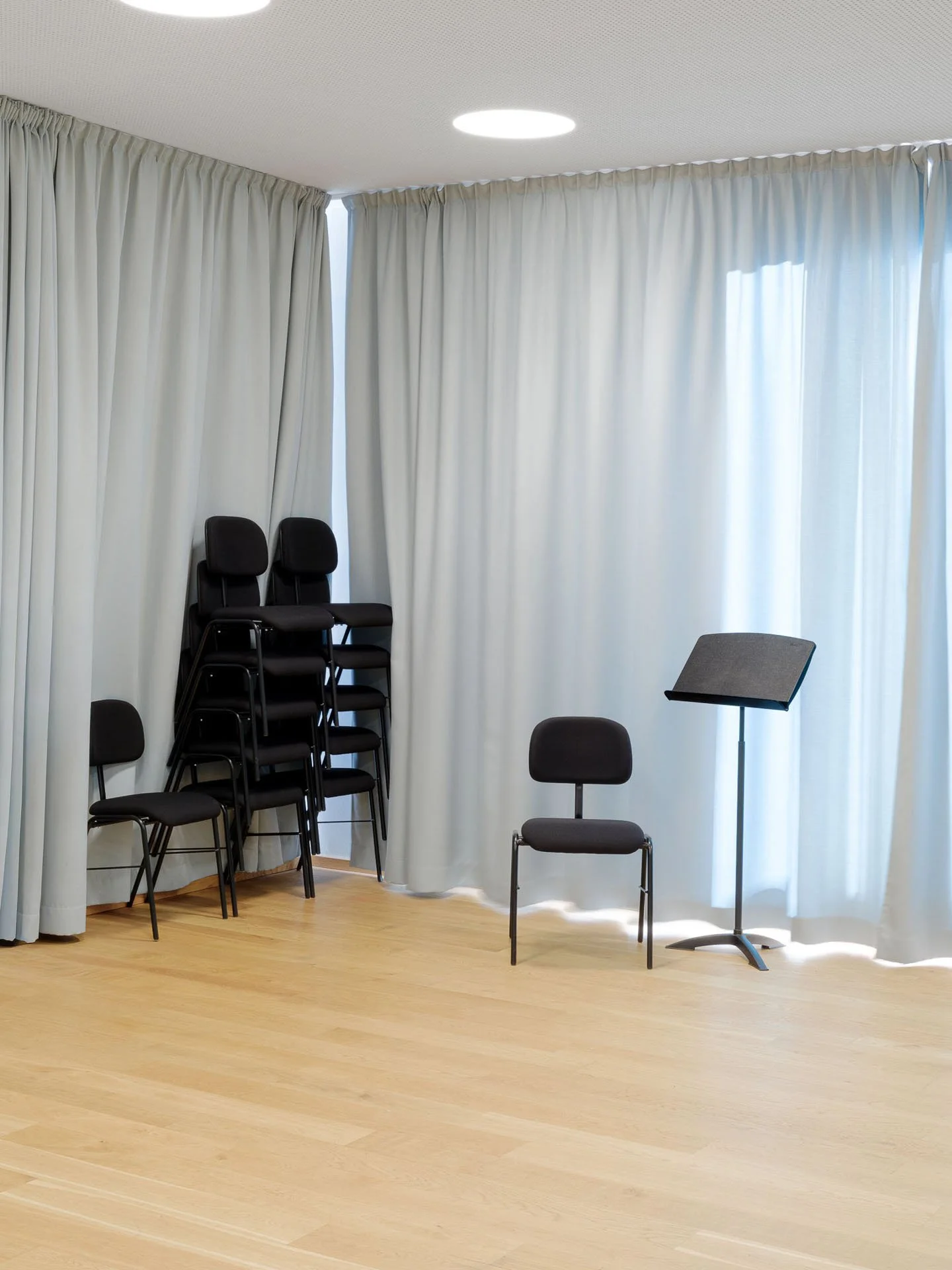 Sala con tende bianche, quattro sedie nere impilate in un angolo e due sedie nere separate al centro, con una copertina musicale di fronte alla finestra.