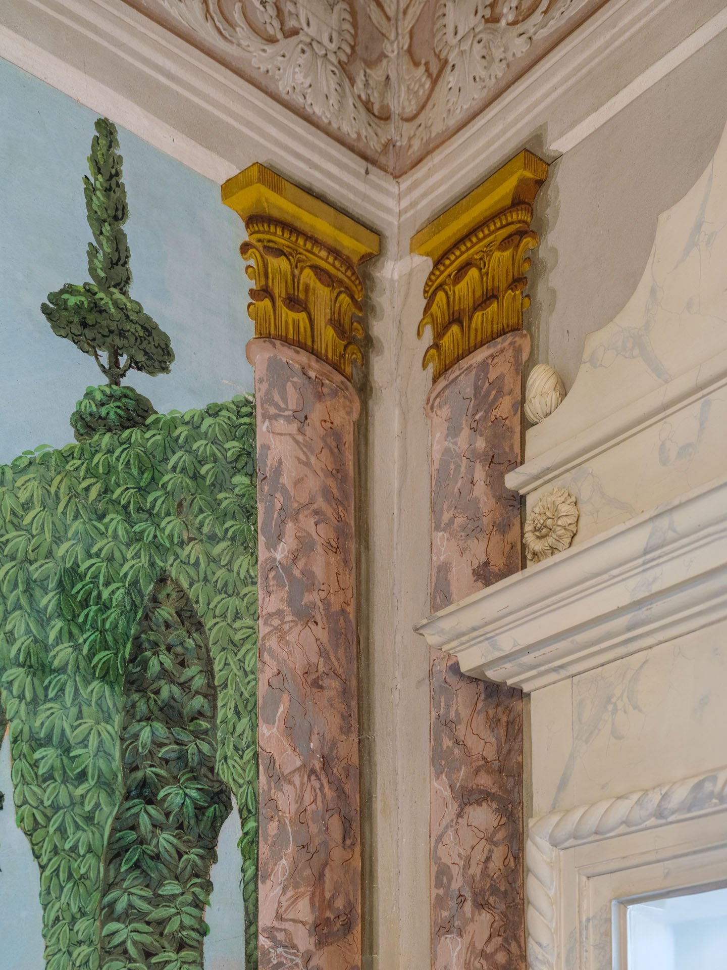 Dettaglio di un angolo di soffitto e parete decorata con stucchi e affresco, raffigurante una scena naturalistica con alberi e rocce, in stile classico.