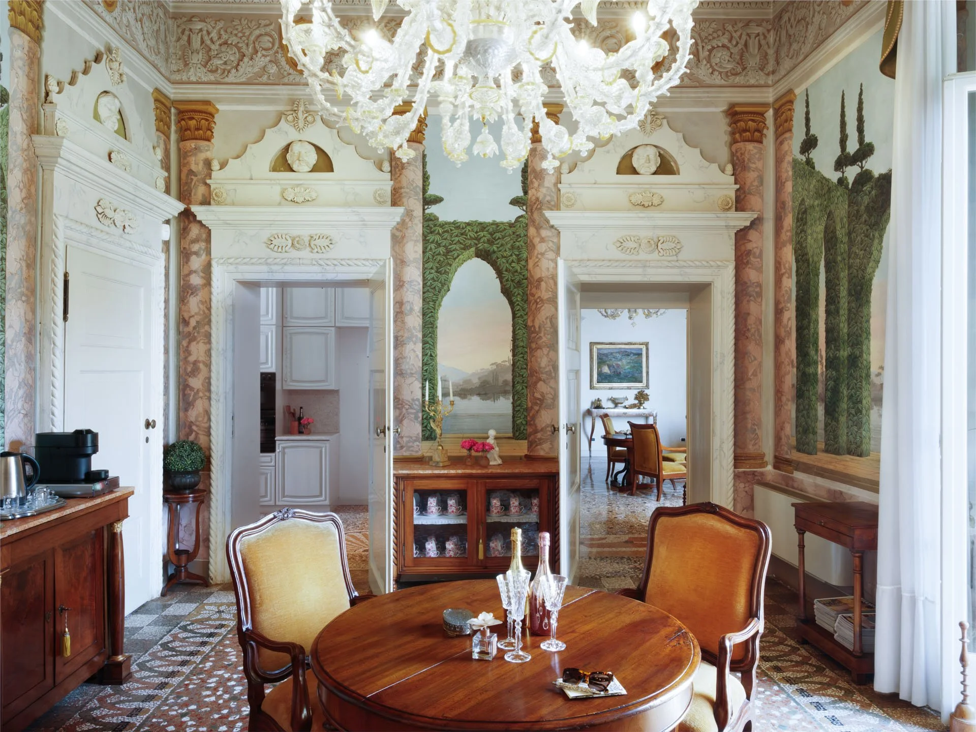 Interno di una stanza elegante con pareti decorate con dipinti e sculture, grande lampadario di cristallo, tavolo rotondo in legno con sedie imbottite, arredi in stile classico, molte piante e quadri alle pareti.