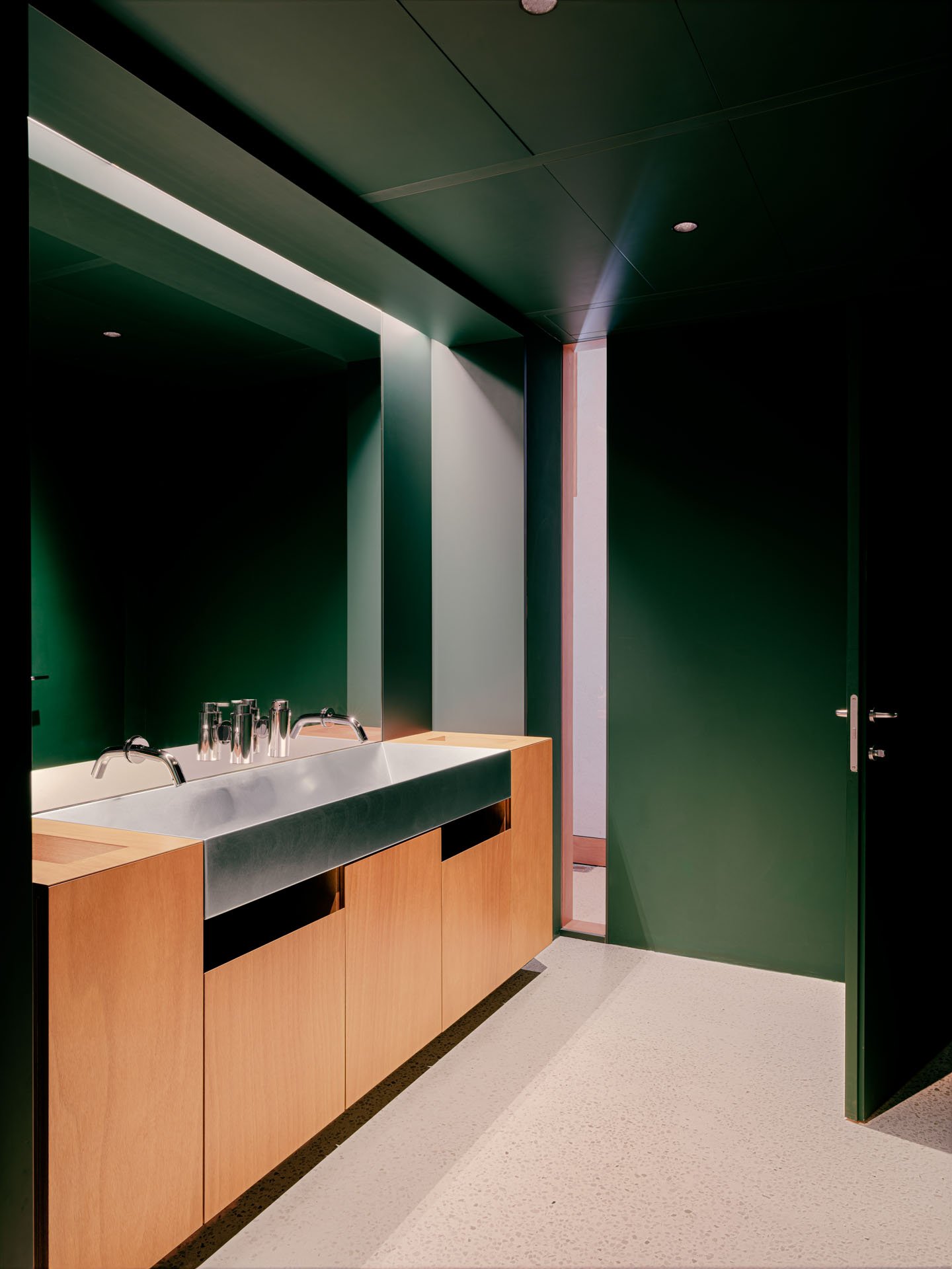 Ripiano con lavabo in acciaio, pareti verdi e porta in legno, ambiente di bagno moderno.