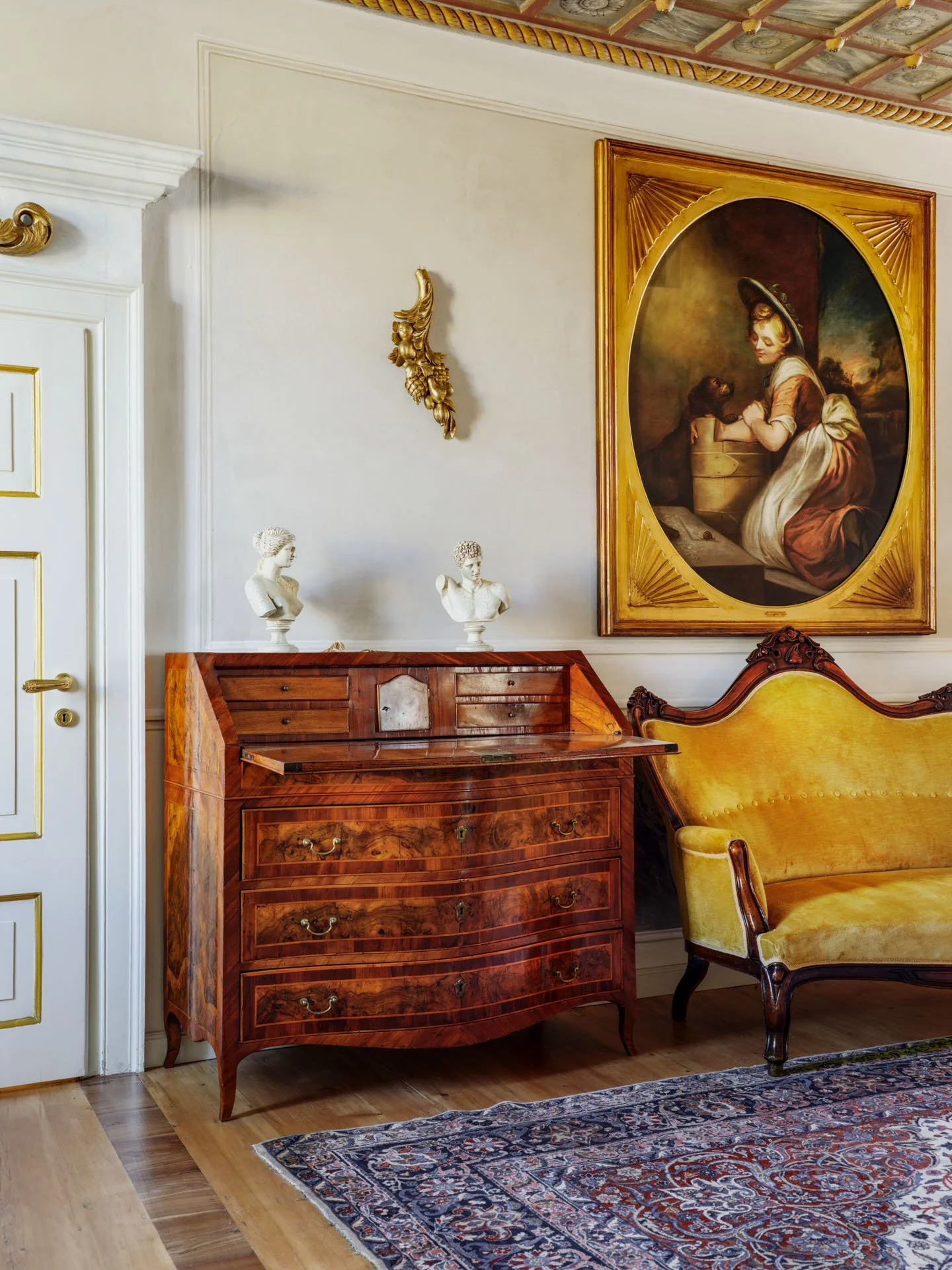 Interno di una stanza con un arredamento elegante, un quadro grande con cornice dorata, un mobile in legno con bustine di marmo e due teste scultoree, un divano in velluto giallo e un tappeto decorato.