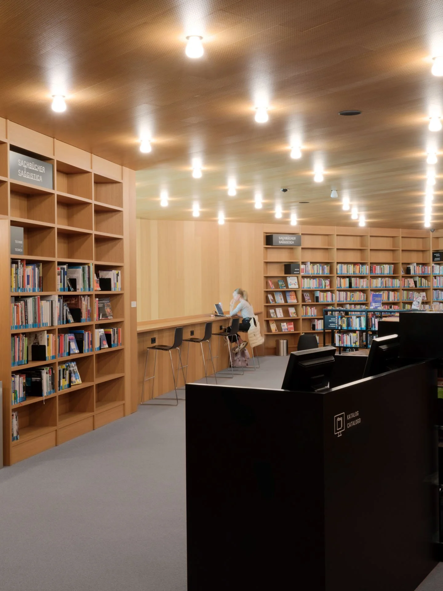Interno di una biblioteca con scaffali di libri di legno e un'area con sedie, alcune persone sedute e che usano il computer.