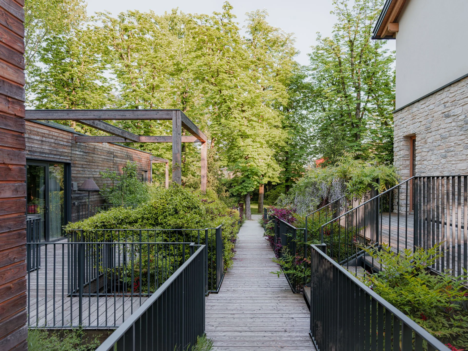 Sentiero di legno tra giardini con piante e arbusti, con case moderne ai lati, e grandi alberi verdi sullo sfondo.