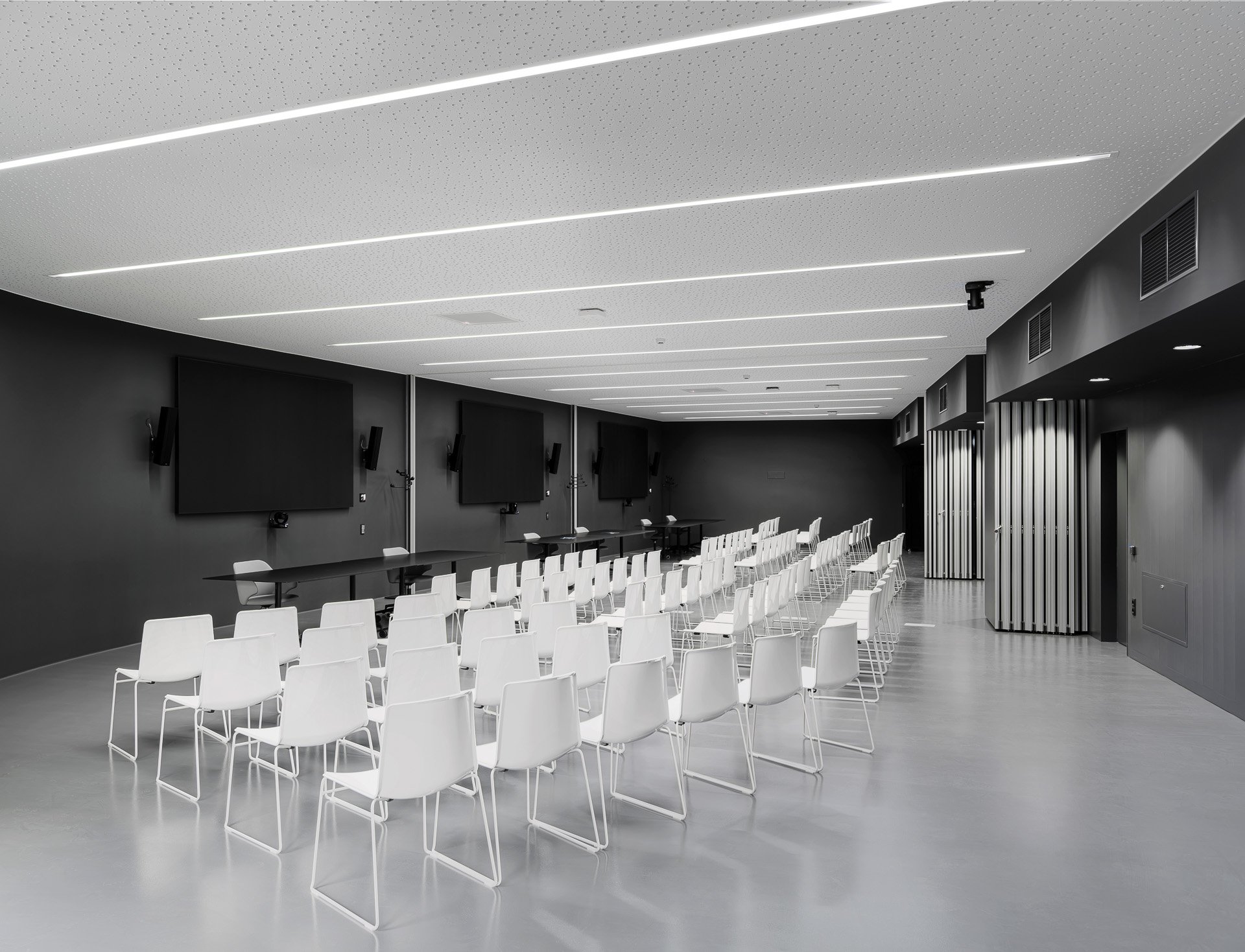Salle conferenze moderna con sedie bianche e grandi schermi neri sulle pareti, illuminata da linee luminose sul soffitto. Il colore predominante è il grigio e il nero.