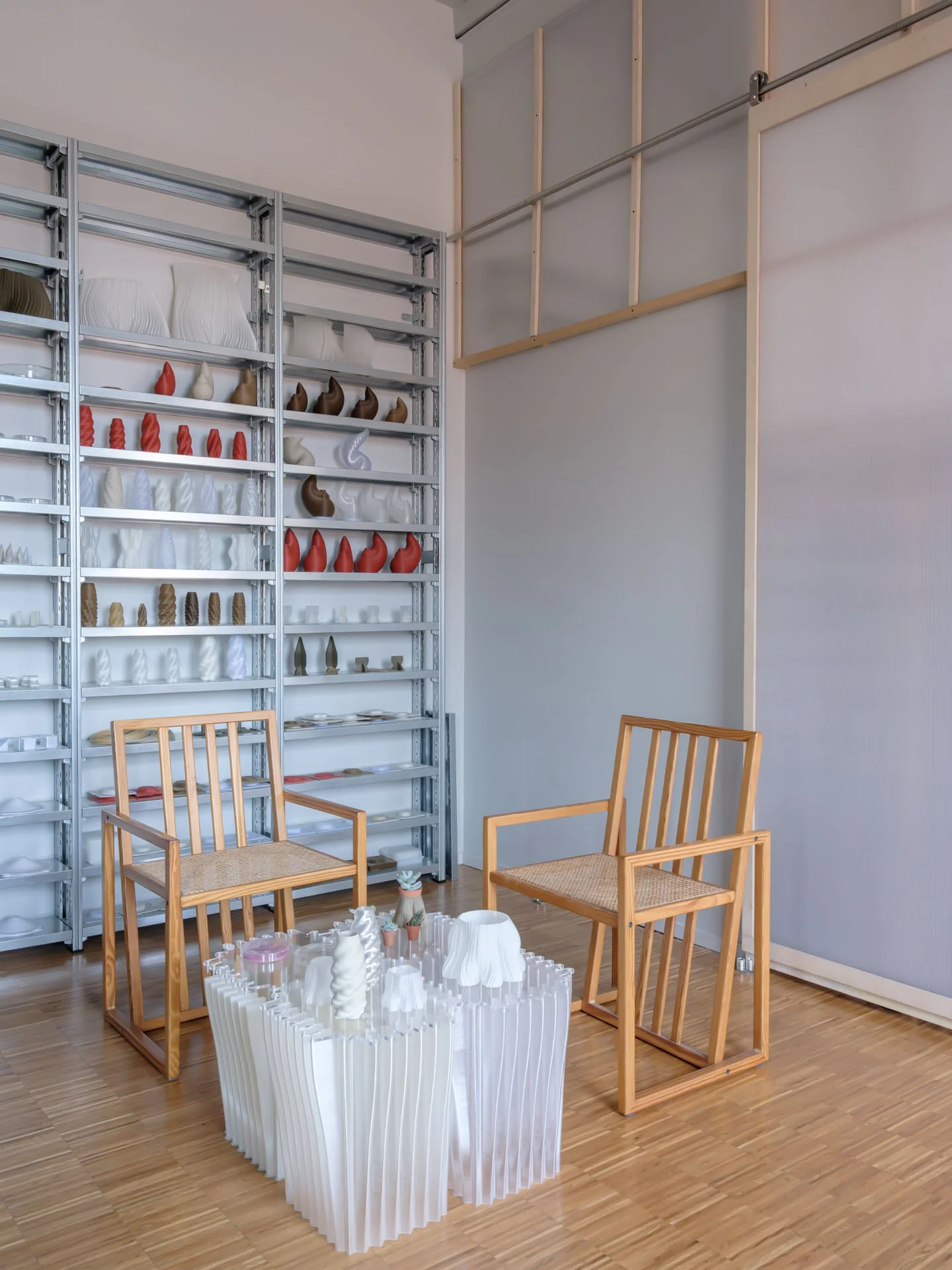Interno di uno studio di design con scaffali pieni di prototipi di oggetti decorativi colorati, due sedie in legno con seduta in paglia e un tavolo ricurvo bianco con oggetti in ceramica.