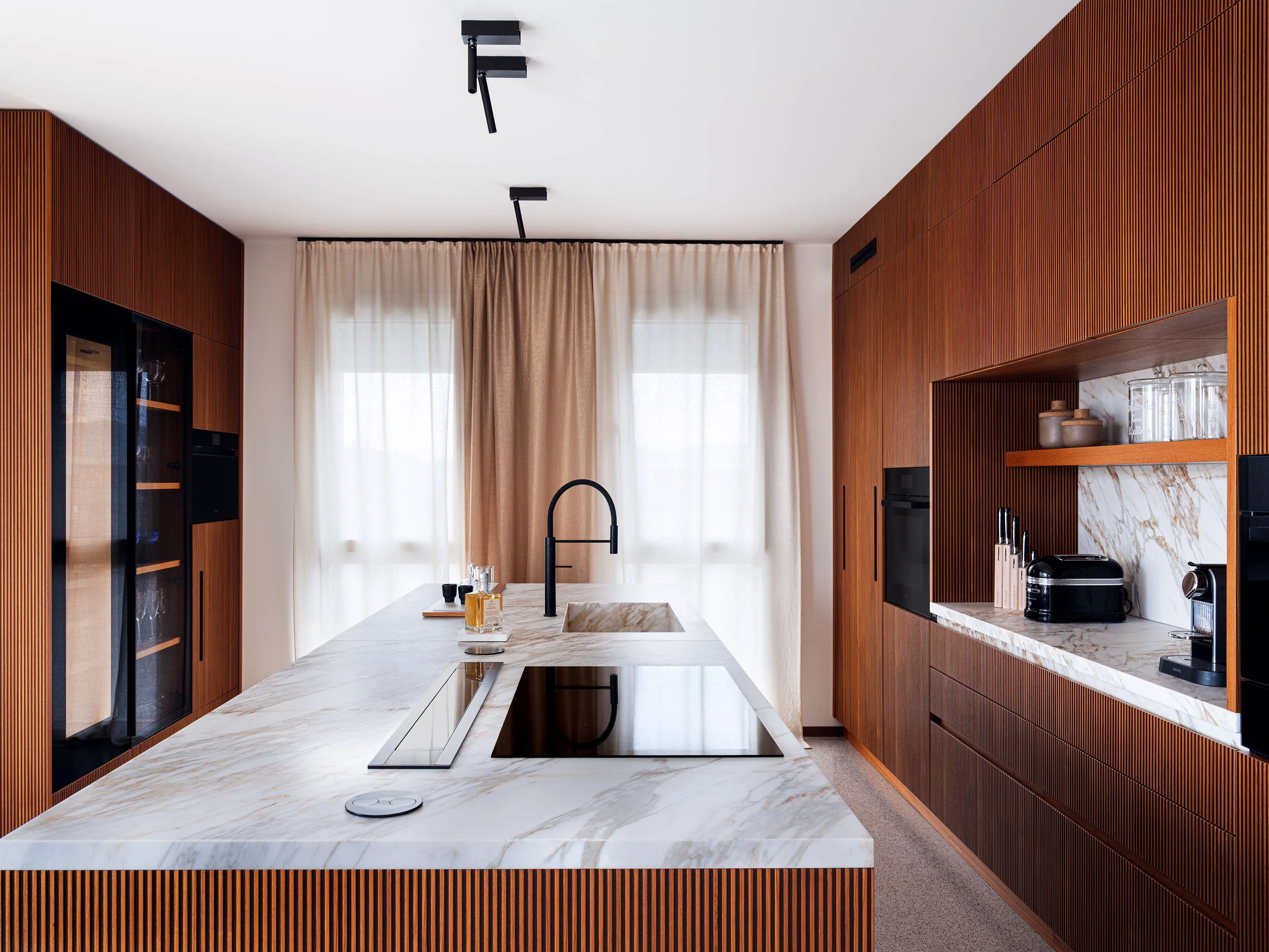 Cucina moderna con mobili in legno e isola con piano in marmo, grandi finestre con tende beige, inizio di due luci nere sul soffitto.