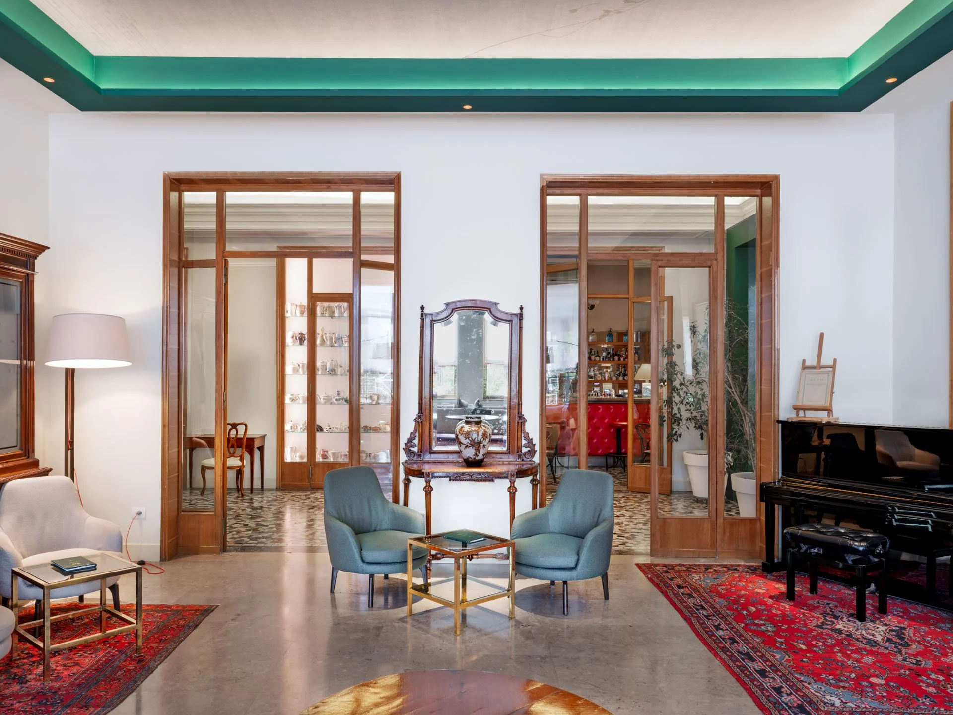 Interno di una stanza con porte in legno che portano a altre stanze, un divano blu, due sedie verdi, tappeti rossi, una richiesta di un pianoforte nero e un grande specchio antico.