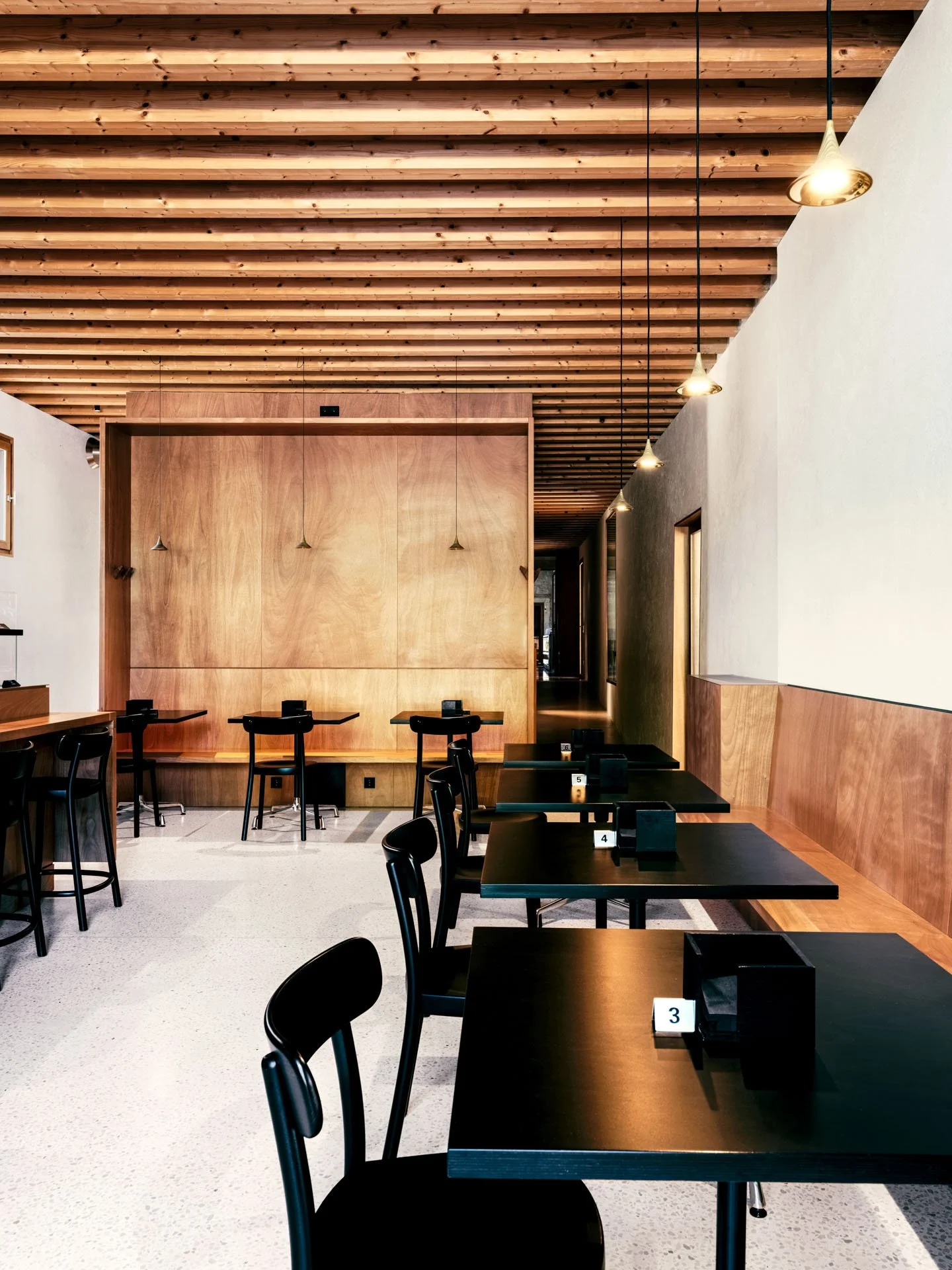 Interno di un ristorante moderno con soffitto in legno, pareti bianche e tavoli in legno scuro, alcuni con segni numerati per servizi. L'illuminazione è data da lampade pendenti dorate. Attorno ai tavoli ci sono sedie nere.
