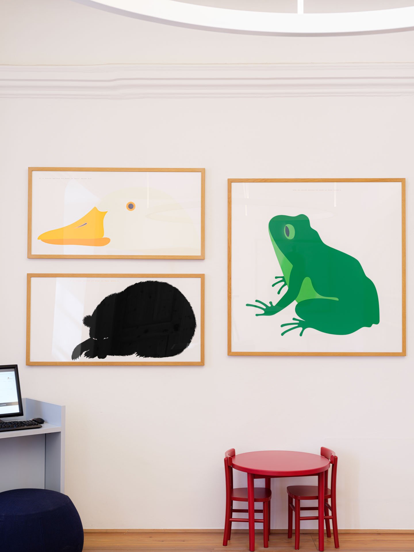 Pareti bianche con tre quadri con disegni di animali: un'anatra, un topo e un coccodrillo, e una piccola area con un tavolo rosso e due sedie, una rossa e una a nido. Sul lato sinistro, c'è una scrivania con un computer.