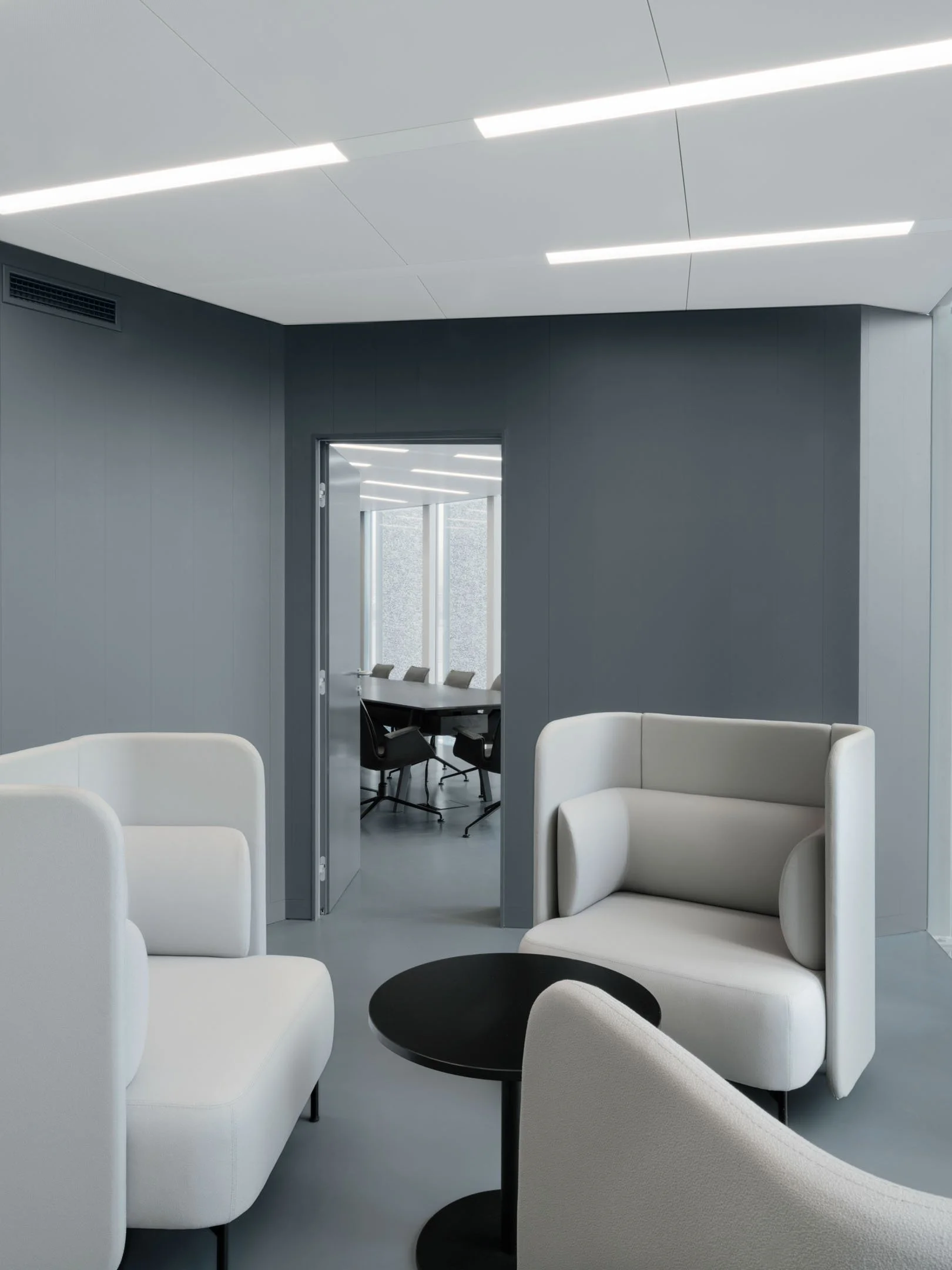 Interno di un edificio moderno con sedie bianche, un tavolo nero, pareti grigie e un soffitto con luci a LED, visibile una sala riunioni con grandi finestre.