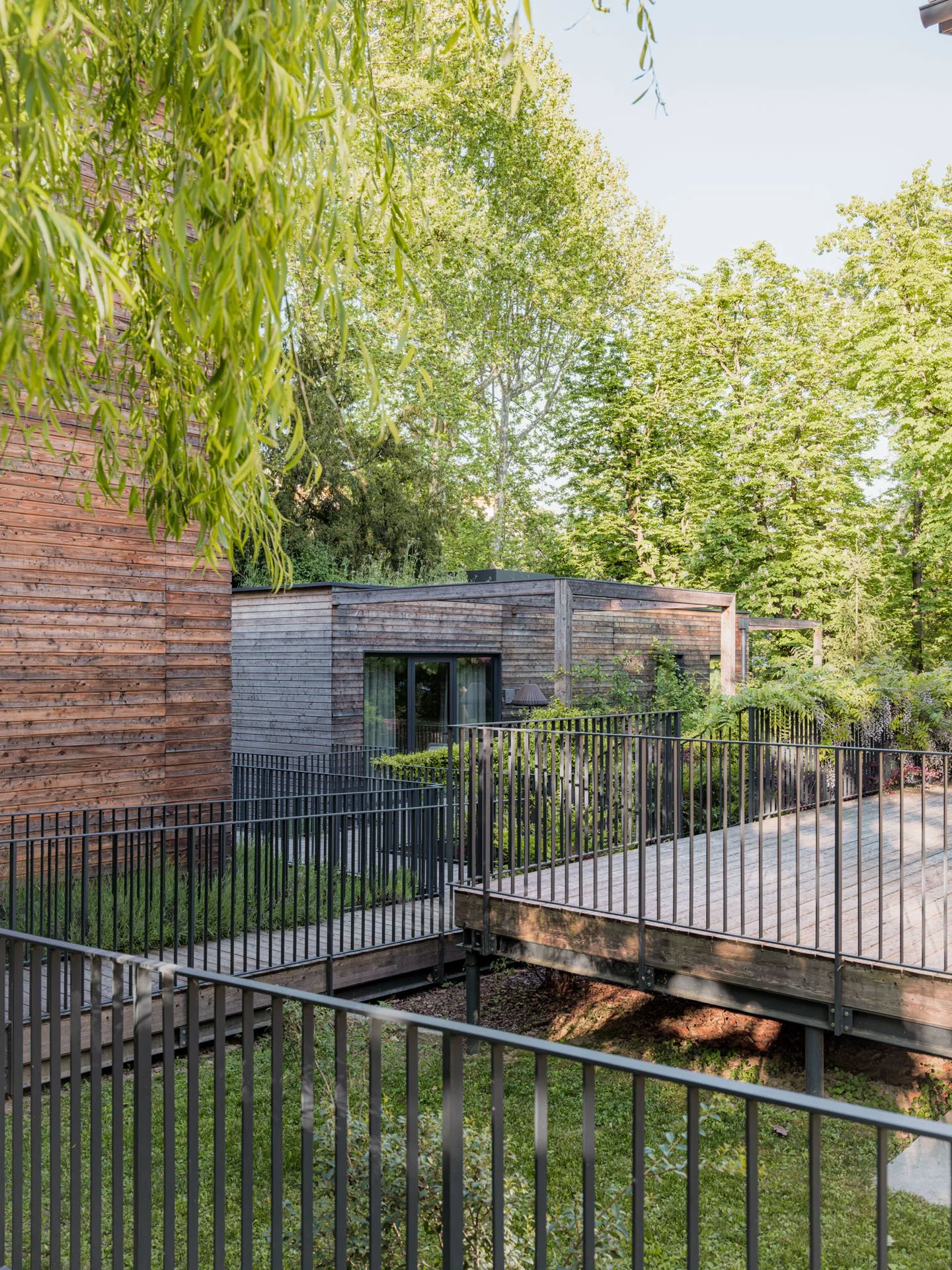 Edificio di sobria architettura in legno con ampio terrazzo circondato da recinzione e immerso in un ambiente verde con molti alberi, in una giornata soleggiata.