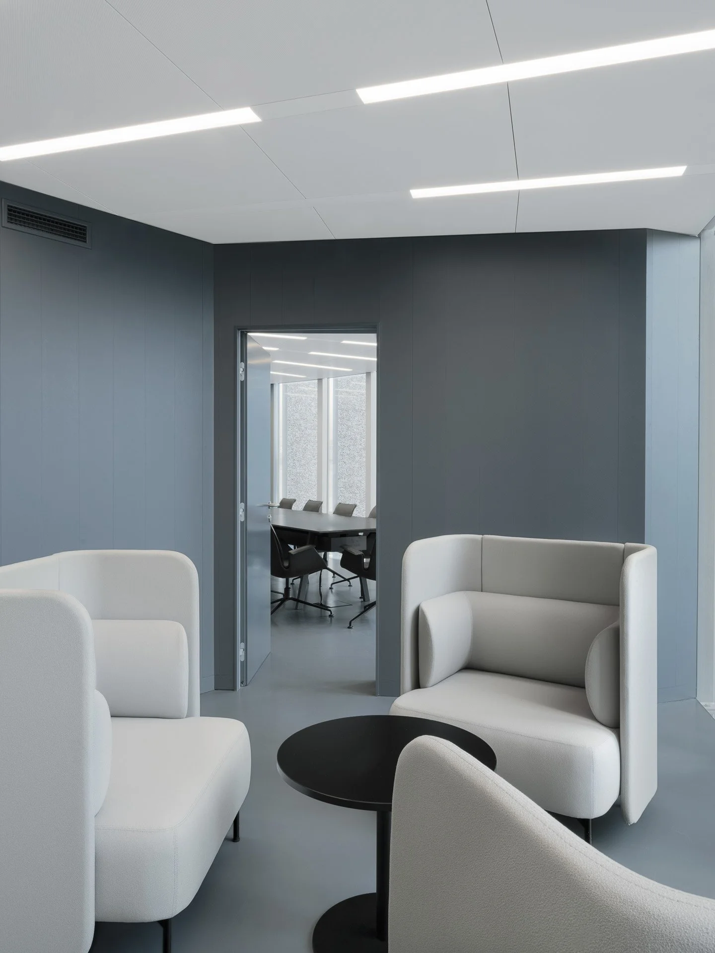 L'ufficio moderno con pareti grigie, sedie bianche e un tavolo bianco, attraverso una porta si vede una sala riunioni con sedie nere e una grande finestra.