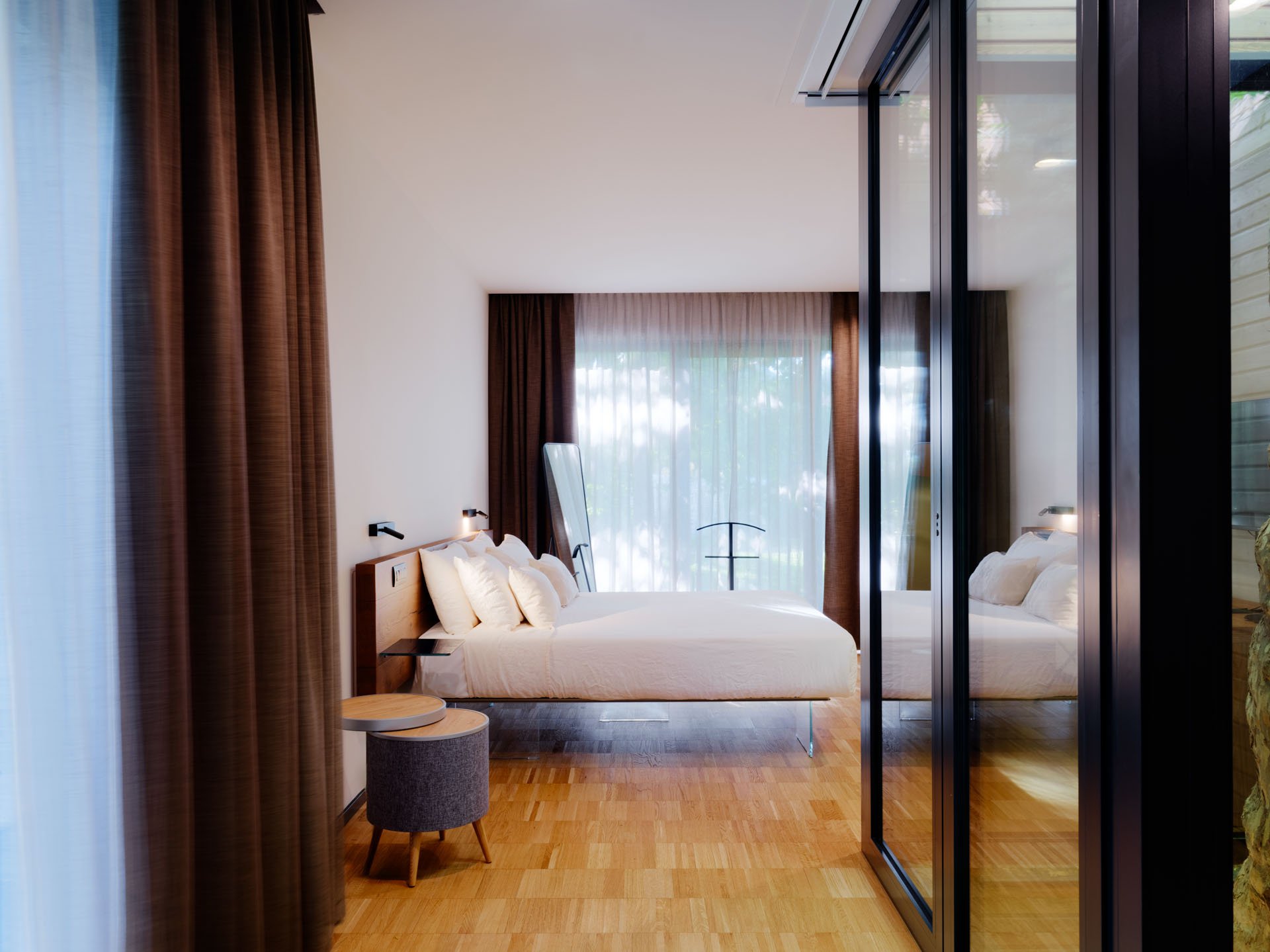 Camera da letto moderna con un letto con testiera in legno, letto con biancheria bianca, due cassettiere rotonde di tessuto grigio, grandi finestre con tende marroni, pareti chiare, un vetro divisorio nero tra la stanza e il bagno.