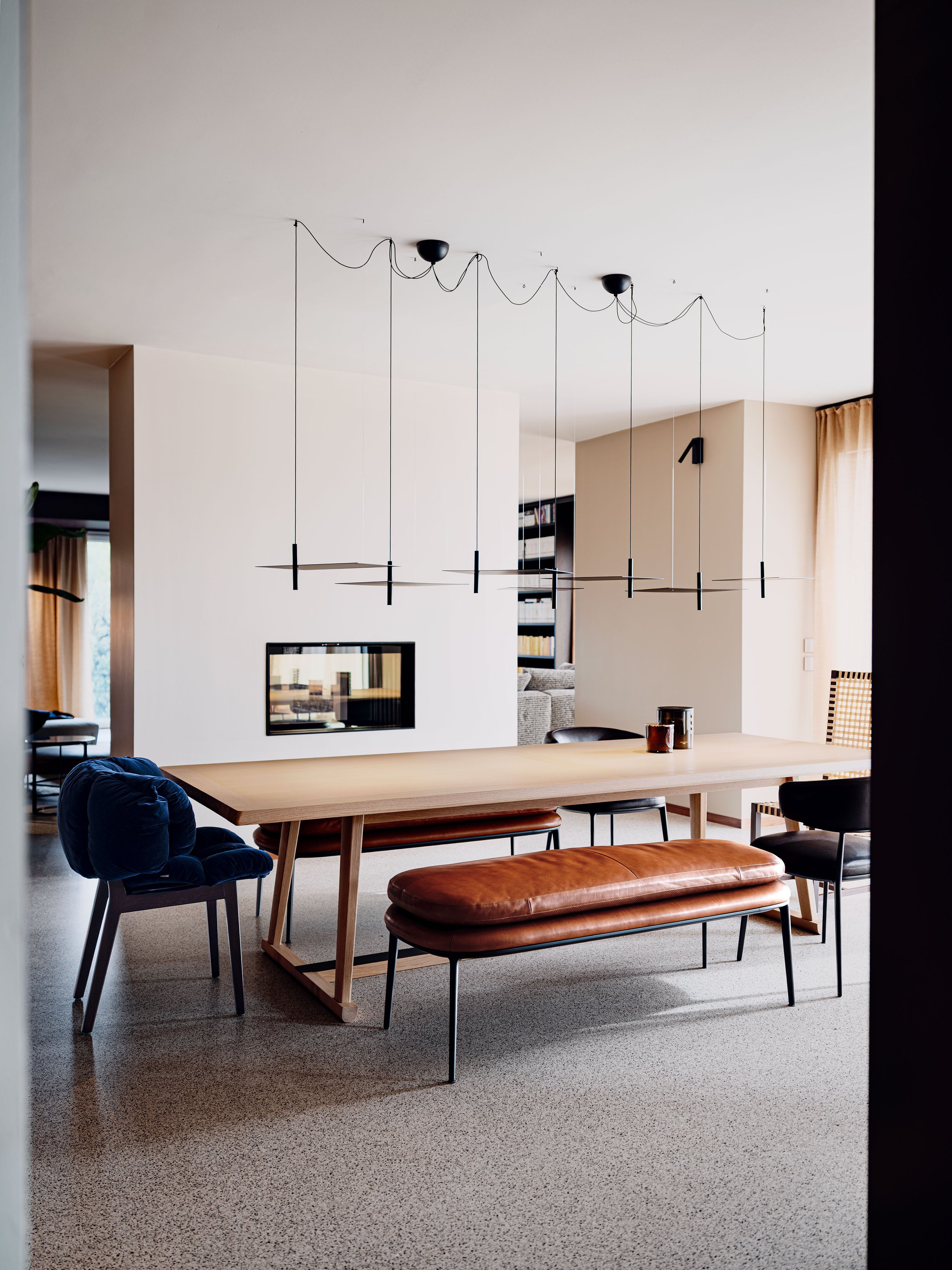Interno moderno con tavolo da pranzo in legno, sedie di diversi stili e colori, lampadario nero sospeso con cavi recisi, pareti beige e una parete con libreria.
