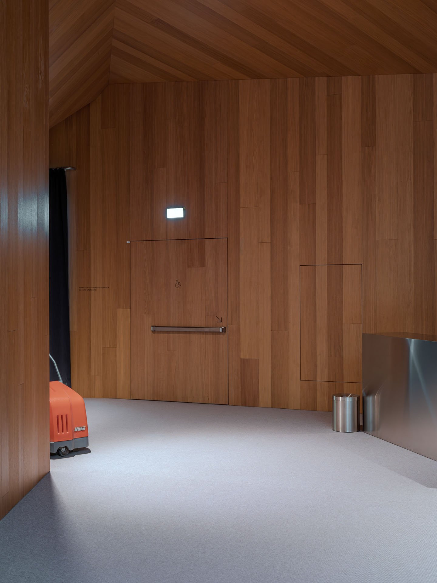 Parete di legno con piccola porta, maniglia e segni di accesso speciale, angolo con cestino e contenitore, su pavimento chiaro.
