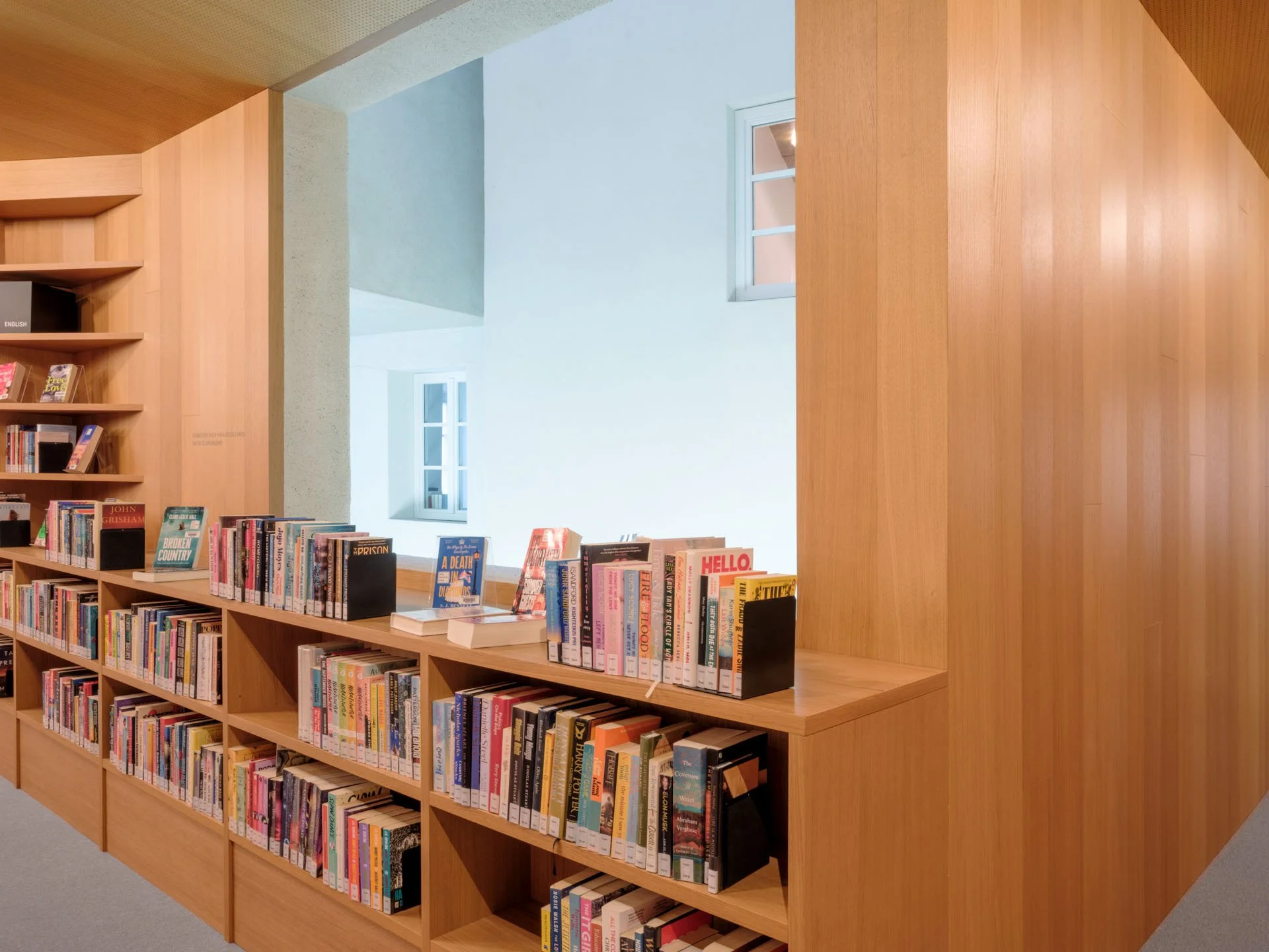 Libreria di legno piena di libri con alcune copie in primo piano, davanti a una finestra bianca su una parete bianca in un ambiente ben illuminato.