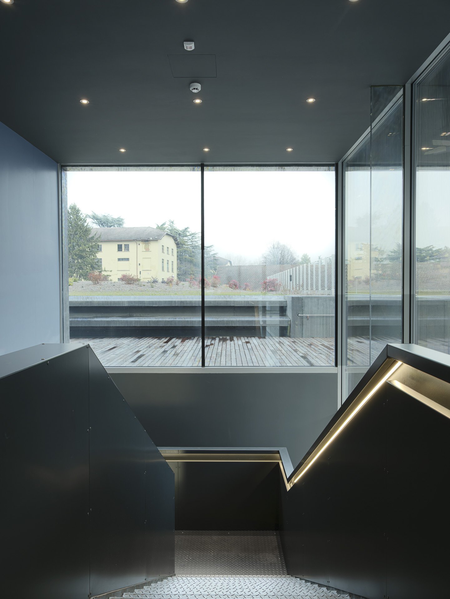 Interno di un edificio moderno con grandi finestre, vista verso l'esterno di case e alberi, con scale di metallo e pareti nere.
