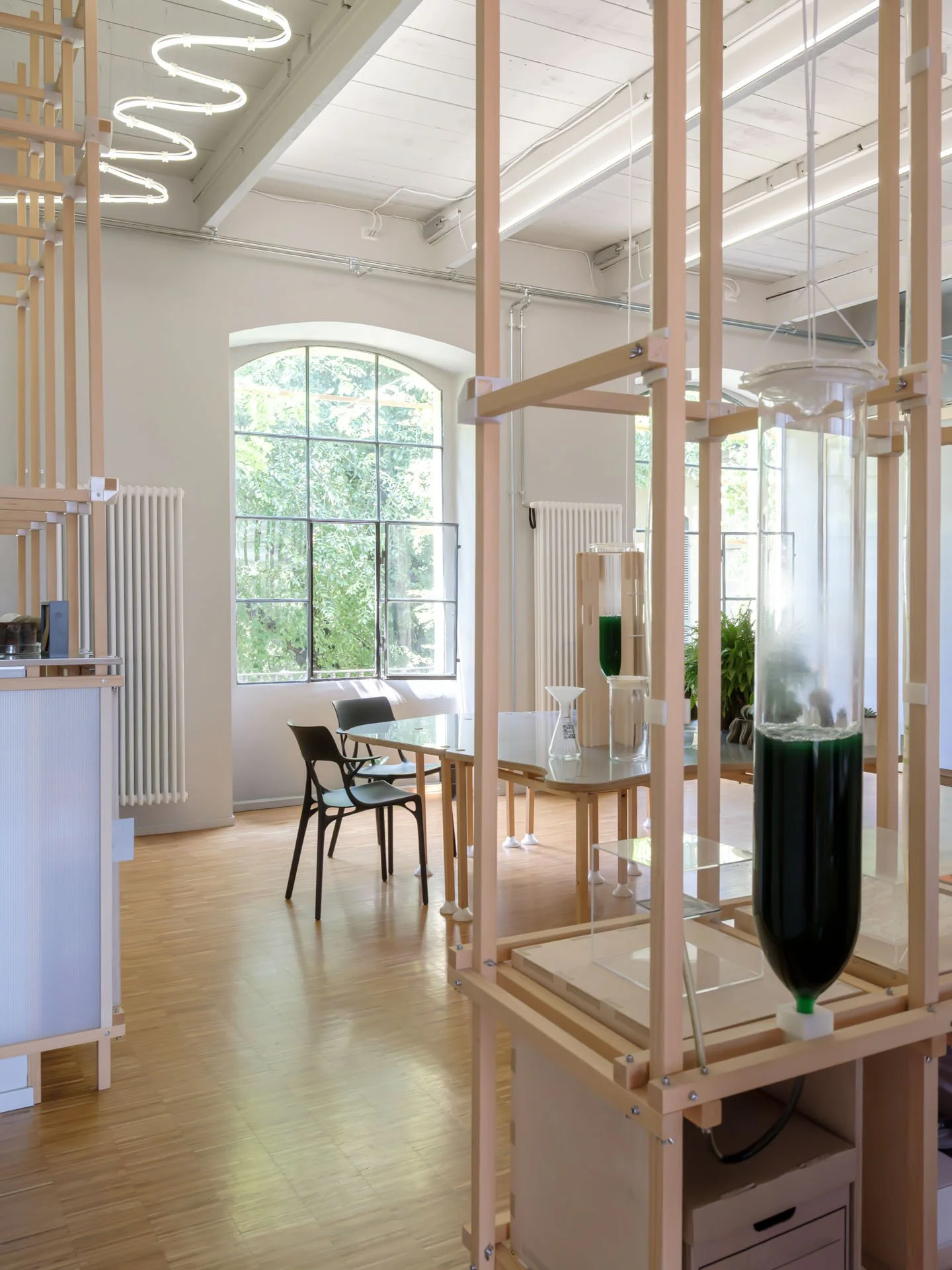 Interno di un laboratorio o aula con grandi finestre, mobili moderni e una struttura in legno con contenitori di liquidi colorati, probabilmente per esperimenti scientifici.