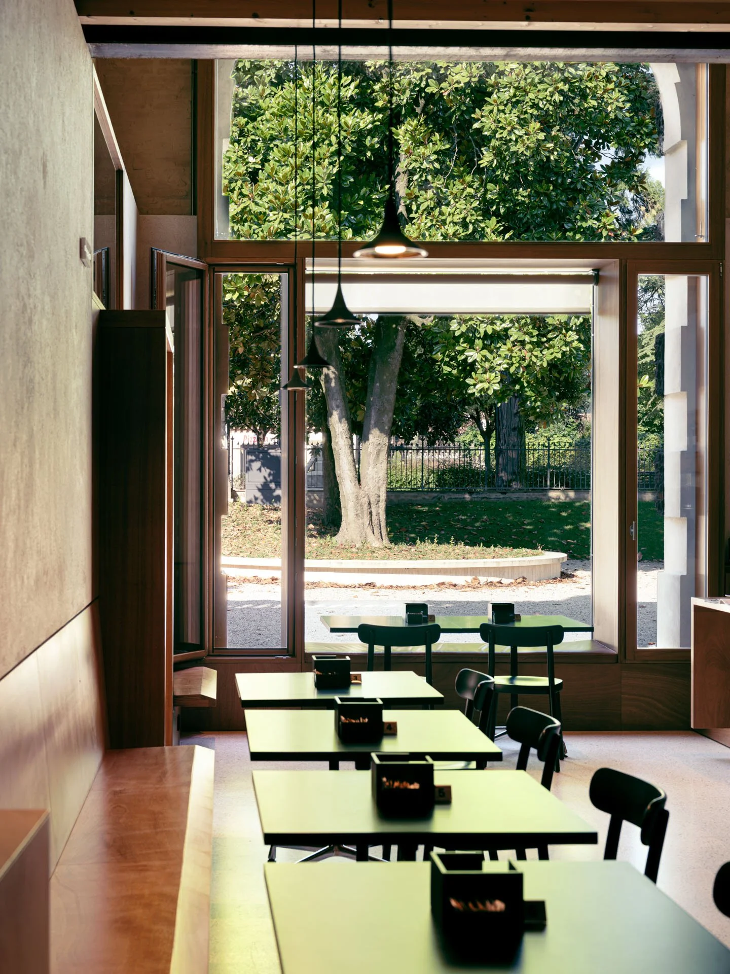Interno di un ristorante con grandi finestra che mostra alberi e natura esterna, tavoli con sedie e luci sospese.