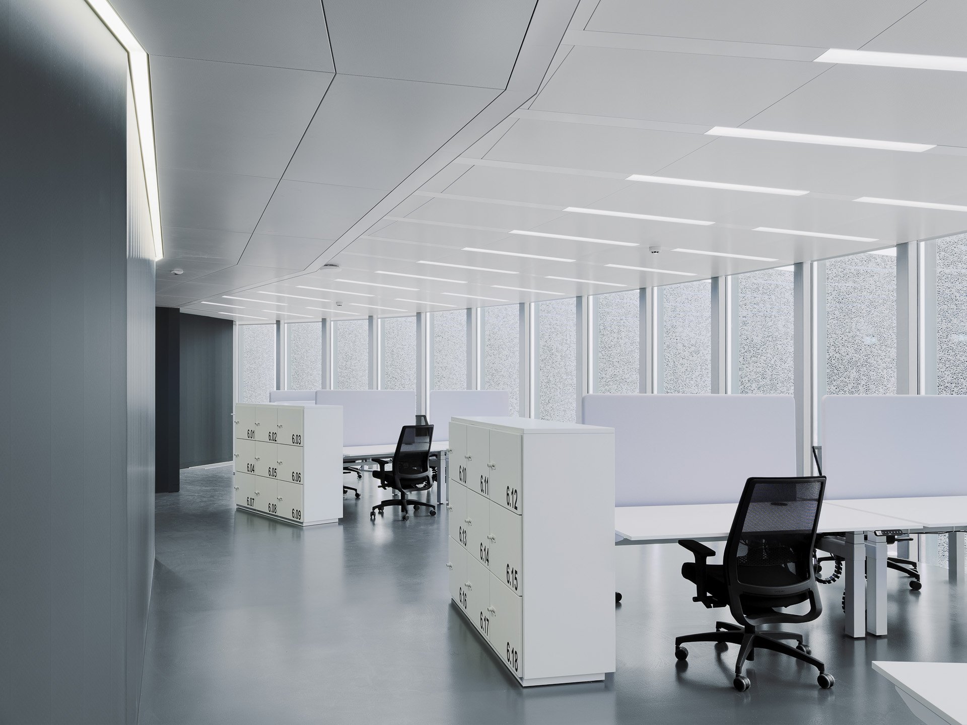 Interno di un ufficio moderno con scrivanie bianche, sedie ergonomiche nere e armadietti con numeri, grandi finestre con vista sul cielo e pareti chiare, illuminato da luci fluorescenti sul soffitto.