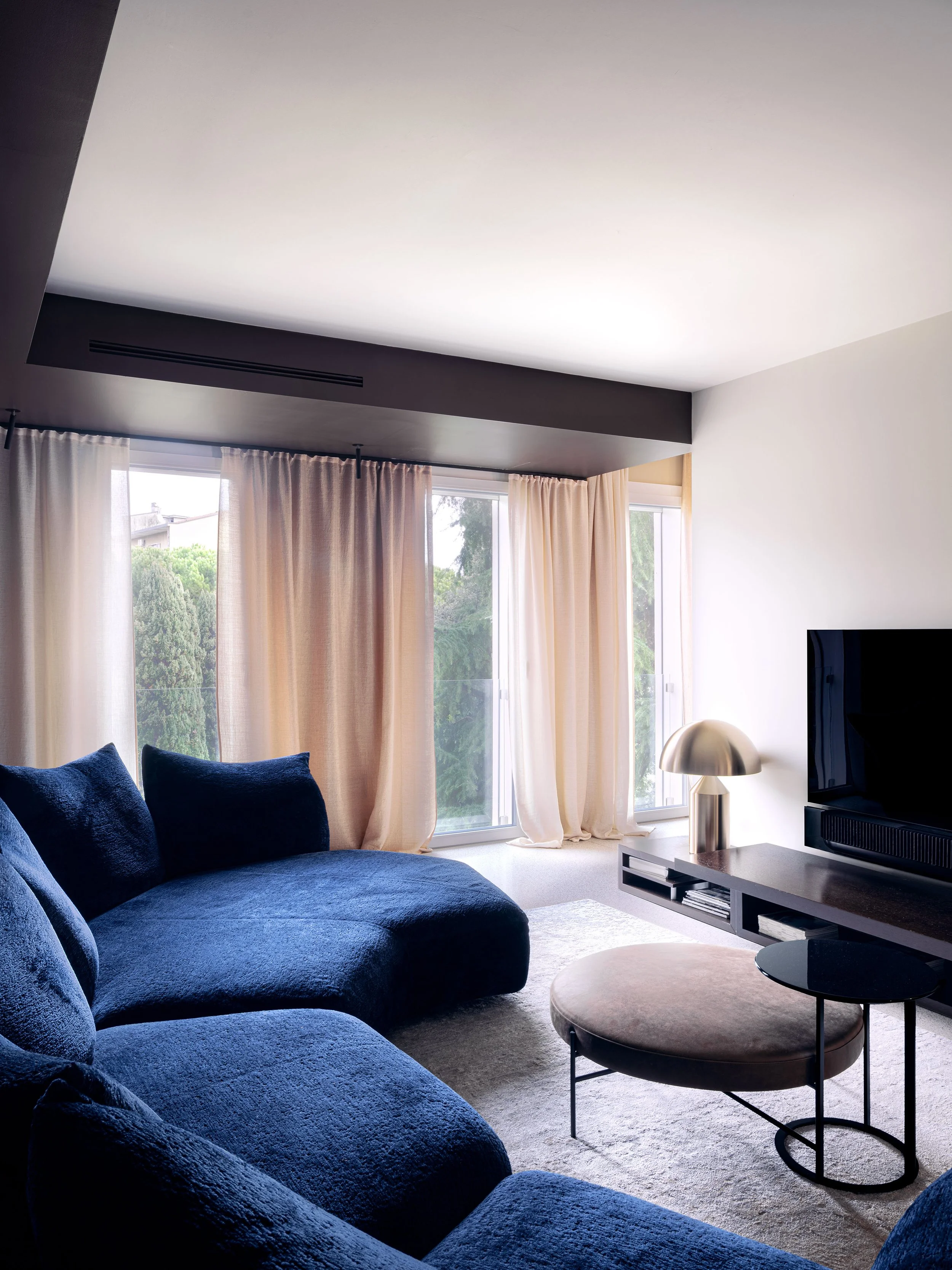 Interno moderno con divano blu, tavolino rotondo con rivestimento beige, lampada a colonna e pareti bianche, grande finestra con tende color crema e vista su alberi e edificio.