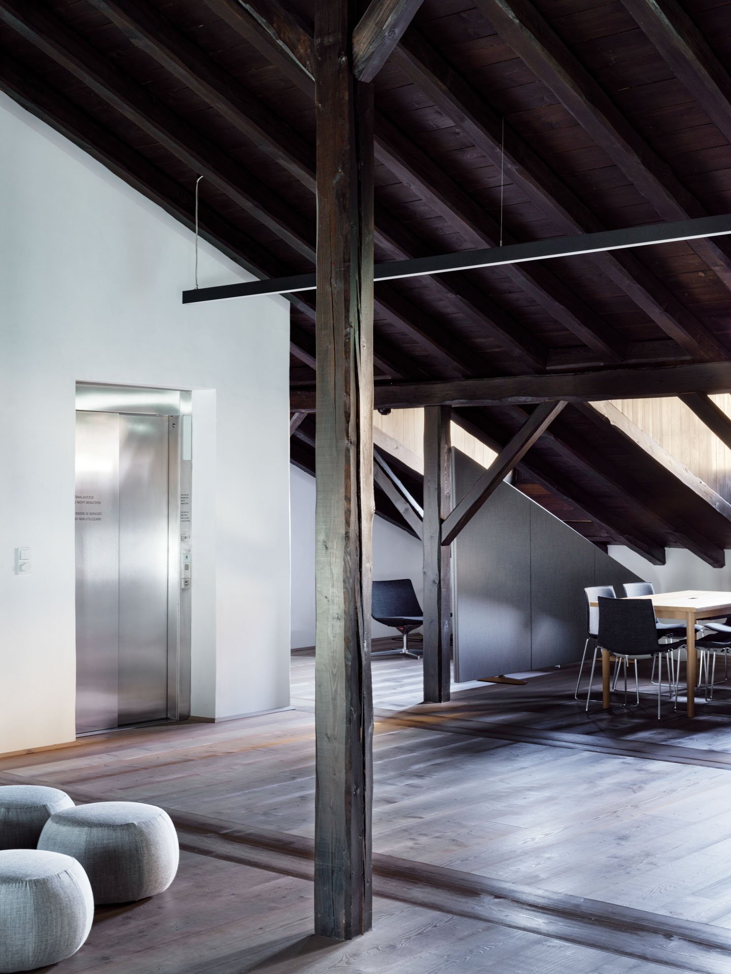 Ambiente interno con soffitto in legno, pareti bianche, ascensore in acciaio, tavoli e sedie, sedute rotondeggianti in tessuto grigio e muretti divisori in moderato stile moderno.