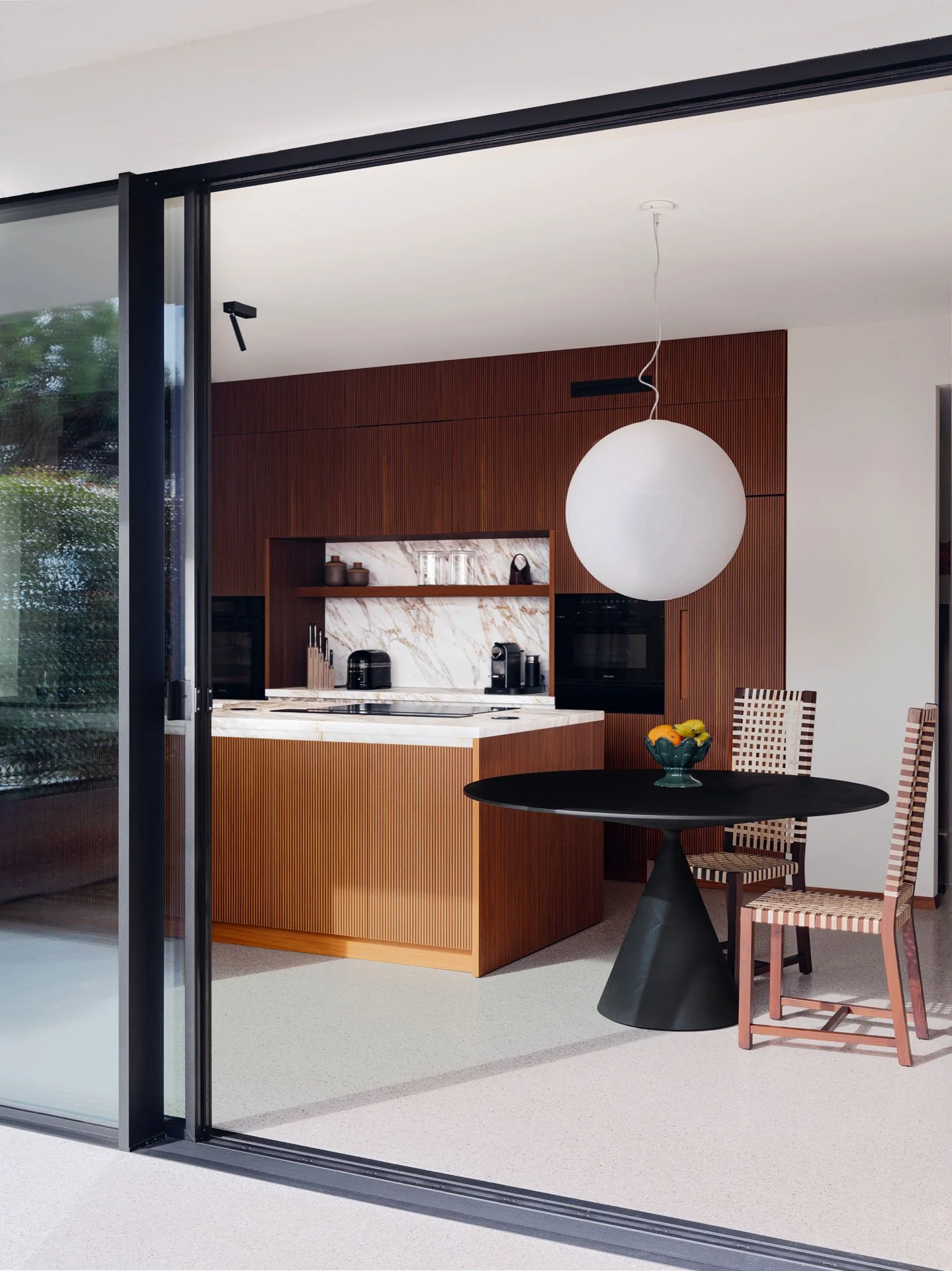 Interno di una cucina moderna con mobili in legno scuro, piano di lavoro in marmo e un grande lampadario sferico bianco. C'è un tavolo nero con due sedie in legno e tessuto a righe rosse e beige, con una ciotola di frutta.