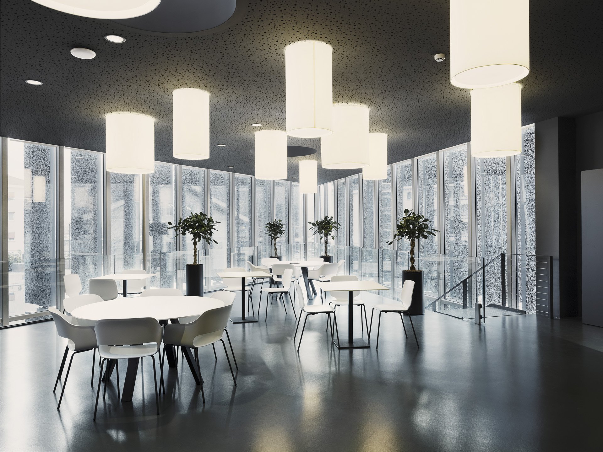 Interno di una moderna area lounge con grandi finestre che affacciano su un edificio, illuminazione a soffitto con grandi lampade e piante decorative.