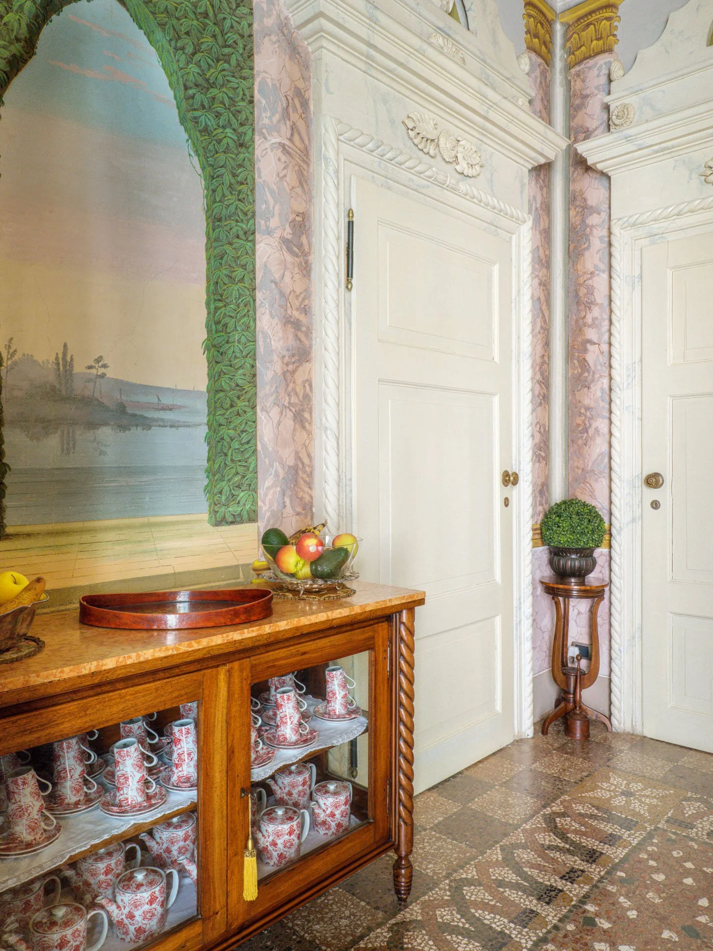 Interno di una stanza con pareti decorate con affresco. Tavolo in legno con vassoio rosso e frutta, vetrina con tazze e piatti decorativi. Porta bianca con ornamenti, vaso di piante su un supporto in legno, e pareti con decorazioni in marmo.