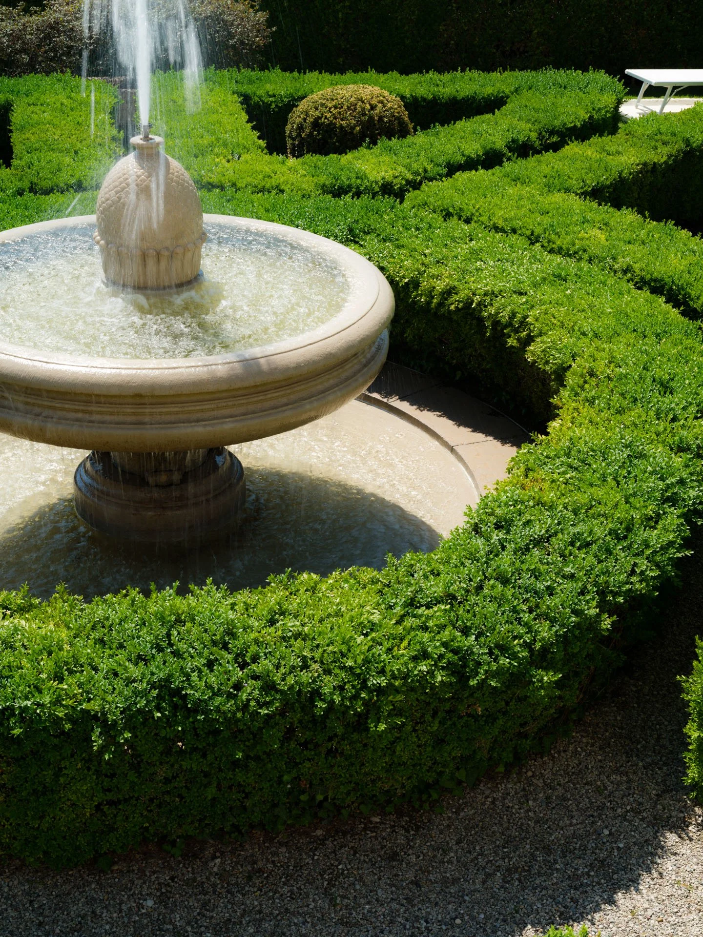 Fontana decorativa con acqua e busto ornamentale, circondata da cespugli verdi ben curati, in un giardino soleggiato.