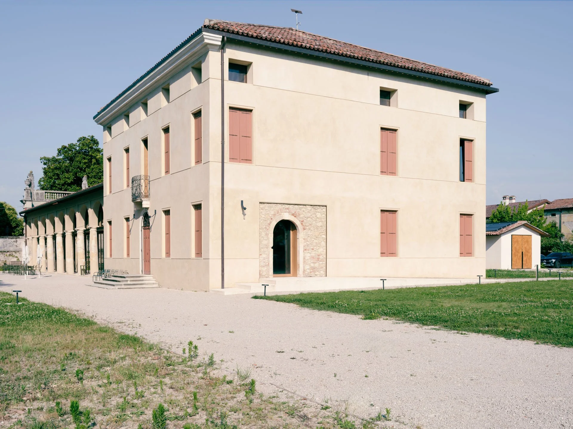 Edificio di tre piani con facciata gialla e finestre con persiane rosa, sotto un tetto rosso, con un'area di prato e un piazzale sterrato in primo piano, al centro.