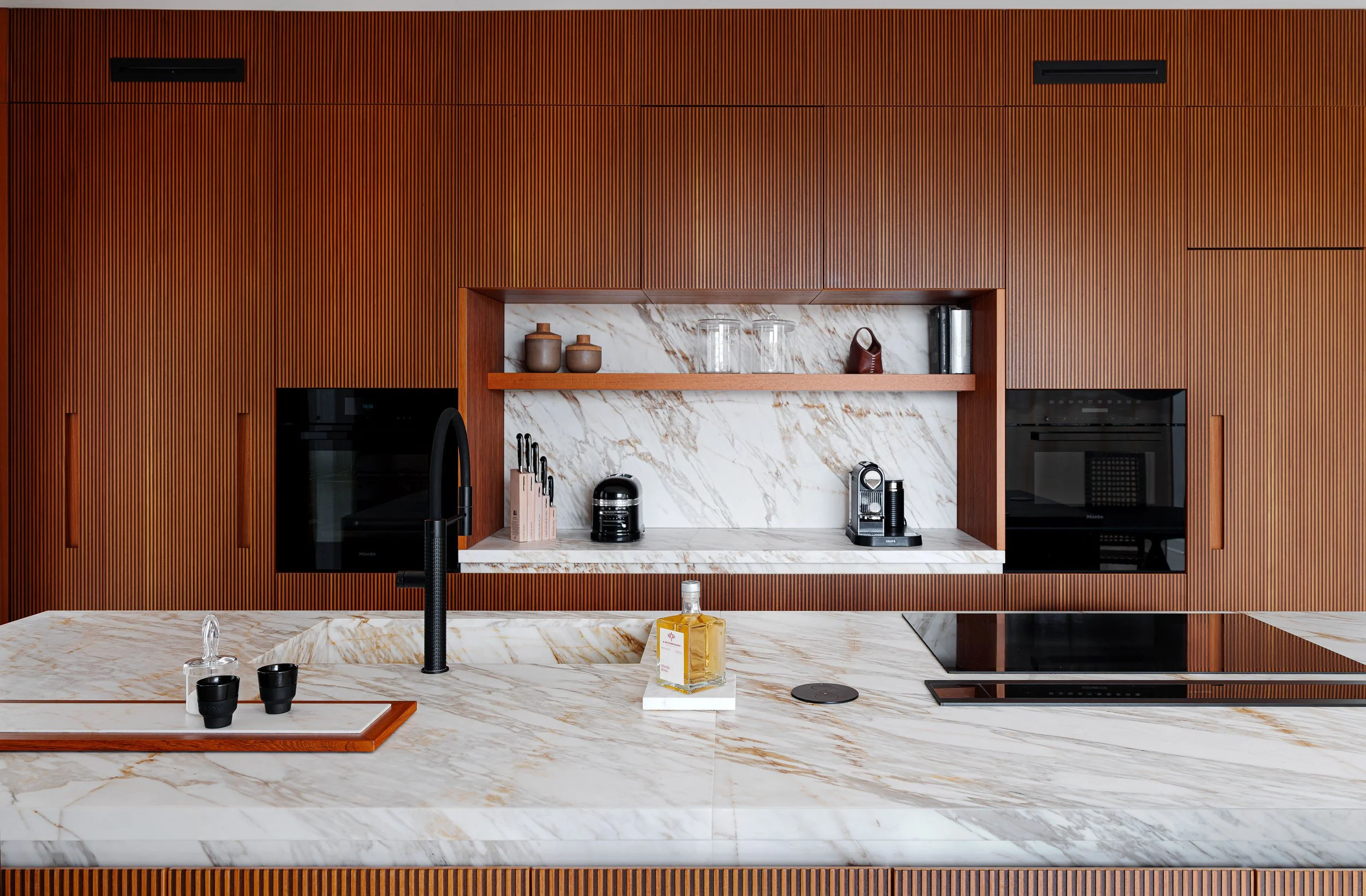 Cucina moderna con ante in legno chiaro verticali, piano di lavoro in marmo bianco con venature dorate, spaziosa e ben organizzata, con oggetti decorativi e elettrodomestici.