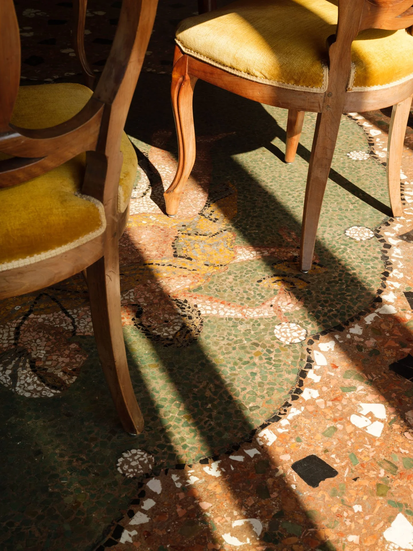 Sedie con sedili imbottiti di velluto giallo e schienali in legno posizionate su un pavimento in mosaico con motivi cerchi e linee colorate.