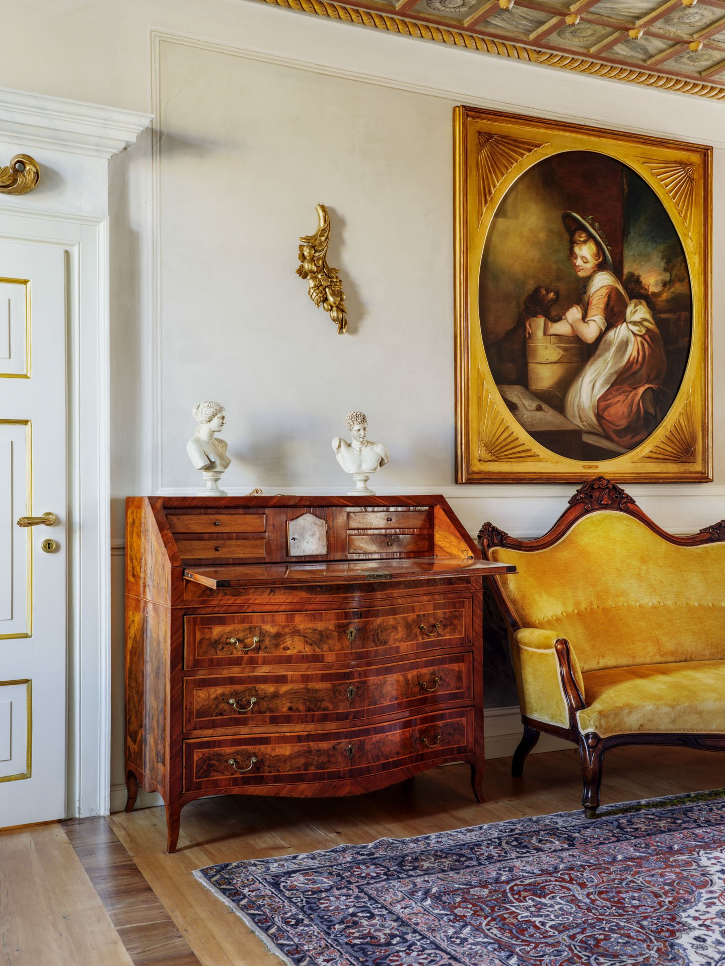 Interno di una stanza elegante con un mobile in legno, due teste di busto in marmo, un quadro con cornice dorata e un divano giallo in stile antico.