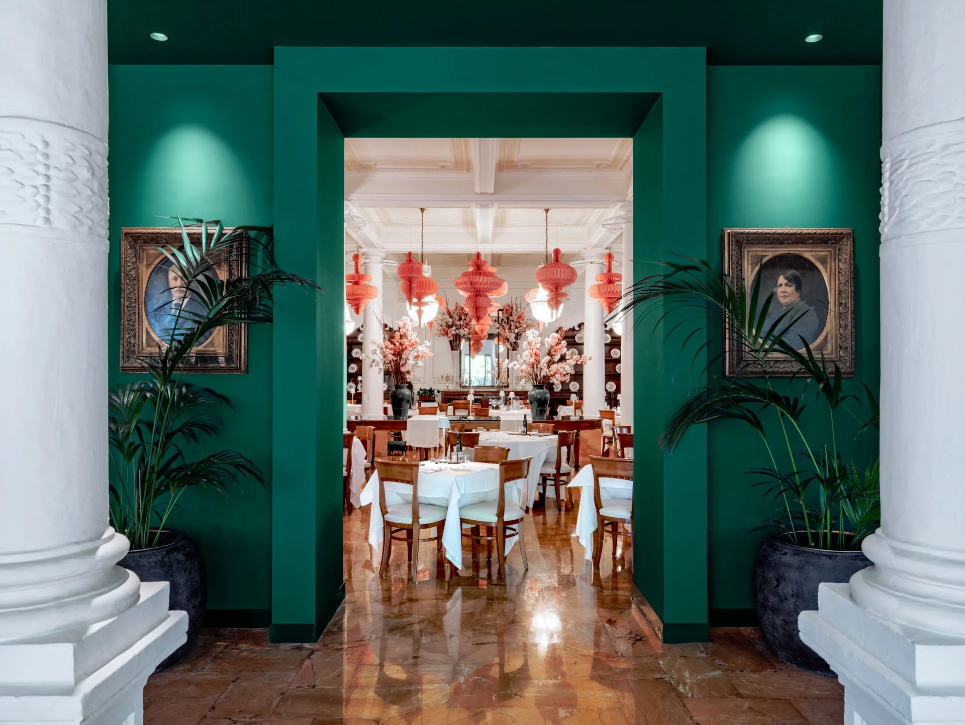 Una sala da pranzo elegante vista attraverso un arco verde, con pareti bianche, dipinti gotici in cornici d'epoca, grandi vasi con piante verdi e un soffitto con lampade di design color rosa.