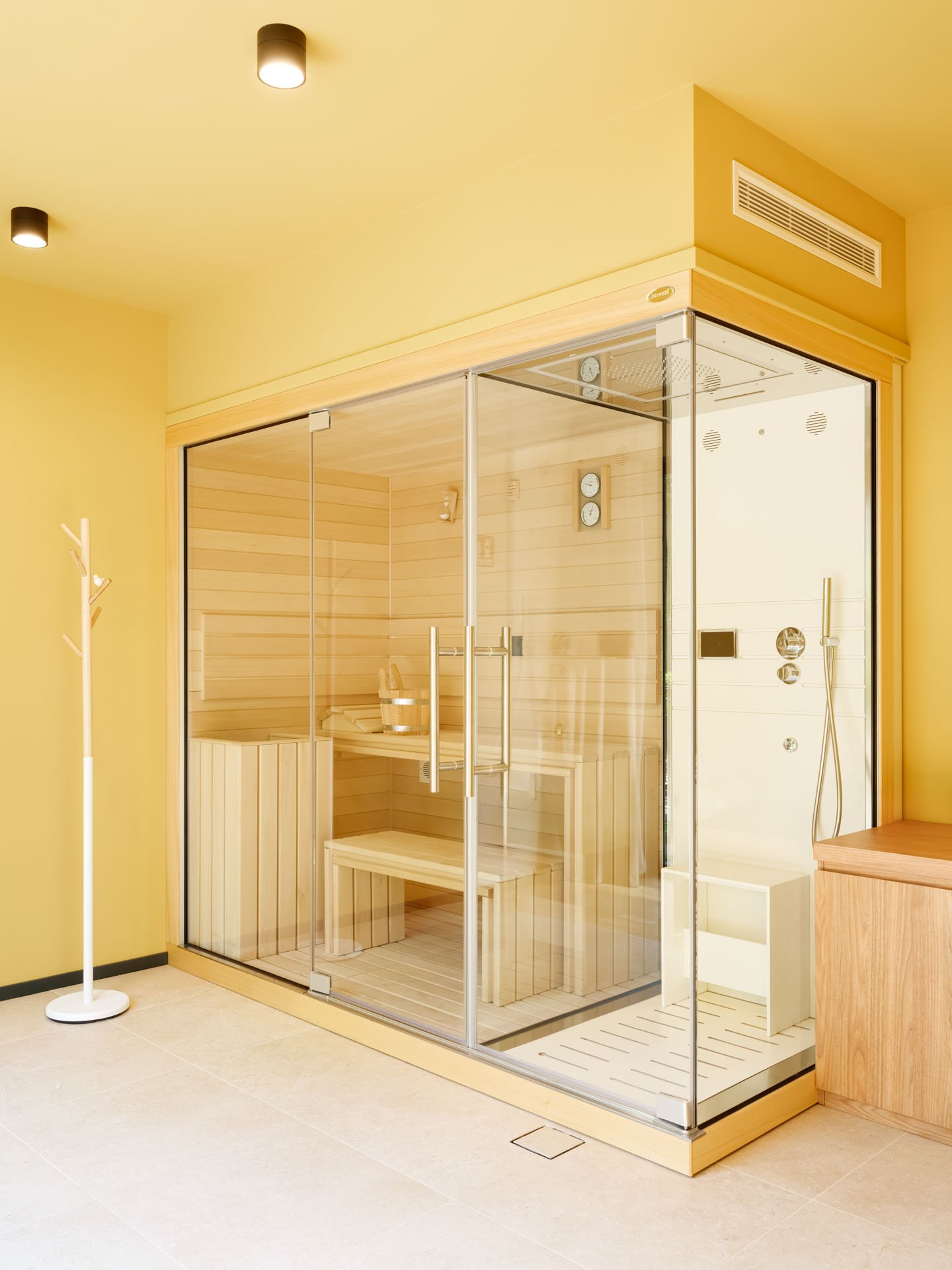 Cabina sauna in legno con pareti di vetro, dotata di sedili e strumenti di controllo, in un ambiente con pareti gialle e pavimento chiaro.