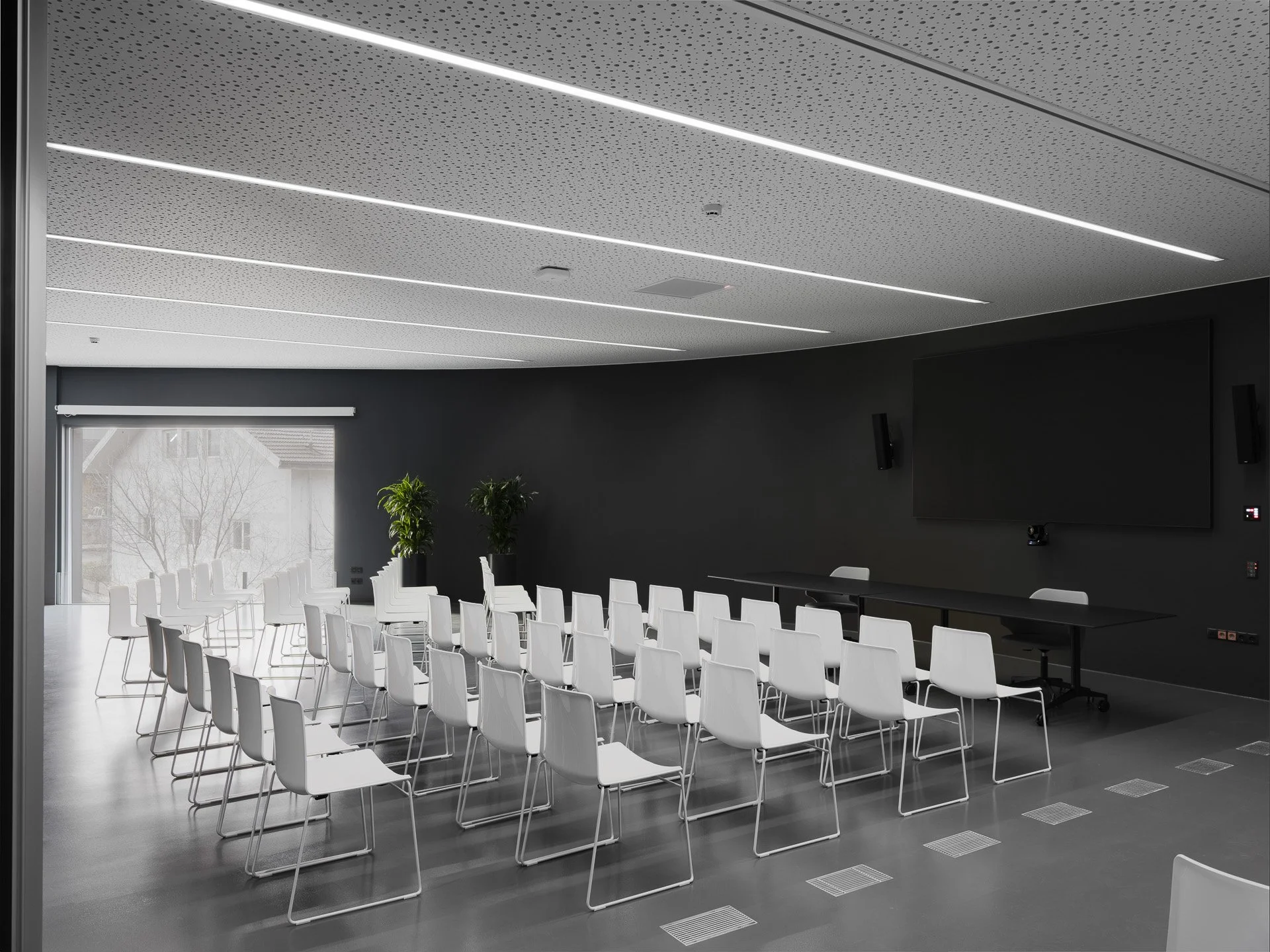 Sala conferenze moderna con sedie bianche disposte in fila, grandi vetrate con vista sul cielo coperto, pareti scure, monitor e telecamere