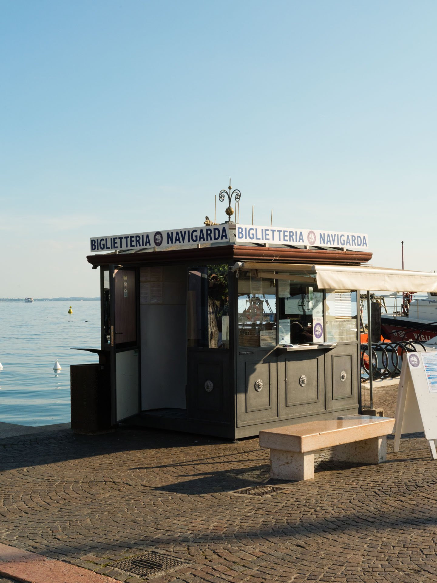 Chiosco per biglietti e collegamenti navali sulla riva di un lago o mare, con un cielo sereno e una barca in lontananza.