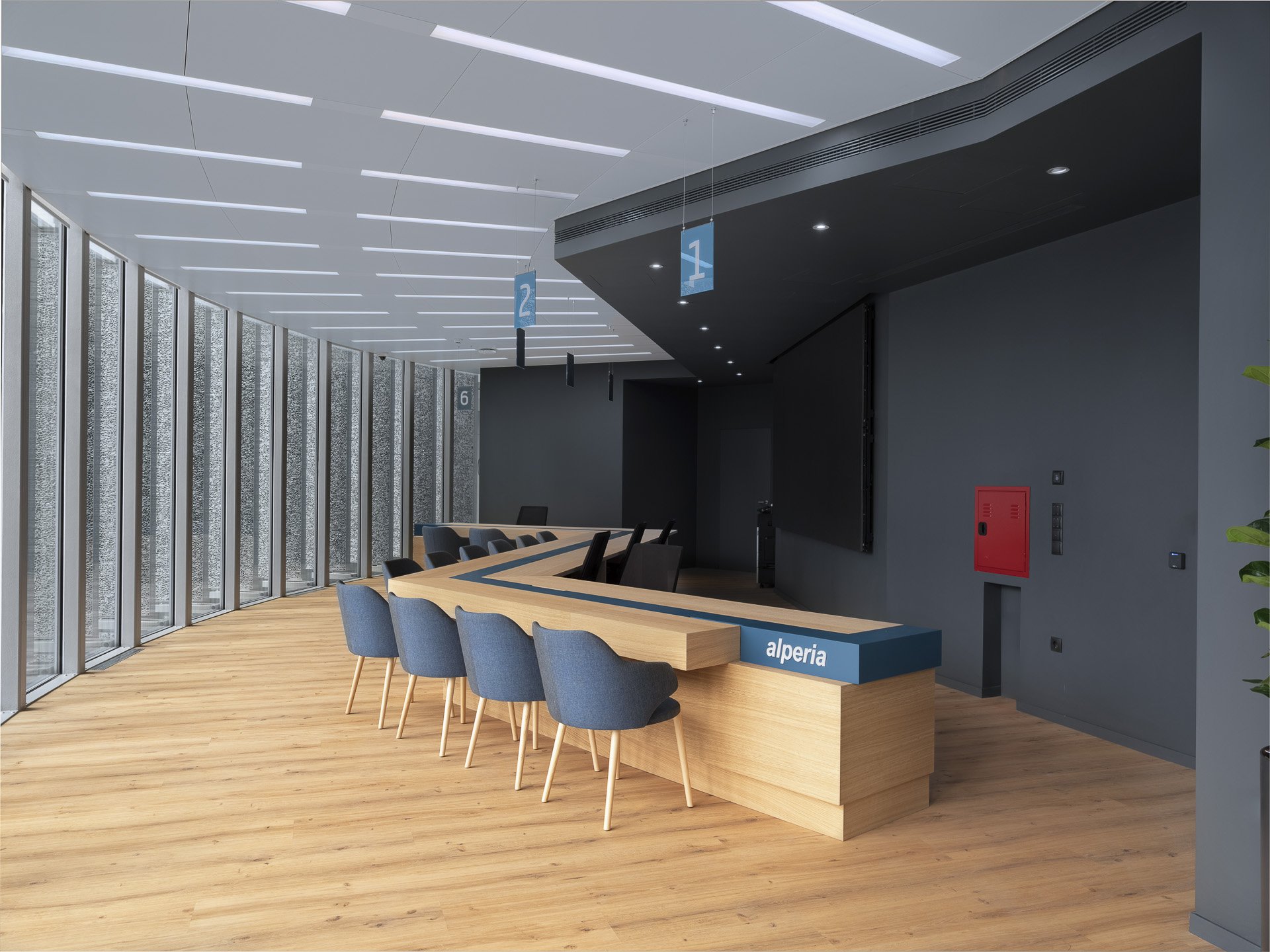Reception area in a modern office with a wooden table, blue chairs, hanging signs numbered 1, 2, 6, and a partitioned glass wall. The ceiling has white linear lights, and there are plants along the side. The desk has the word 'alperia' on it.
